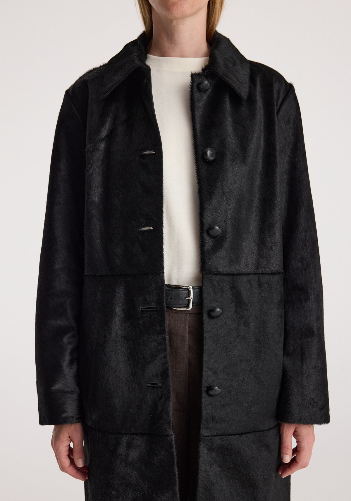 Close-up front view of a person wearing a sleek black ponyhair coat | black with a button-up front over a white top and dark pants with a black belt.