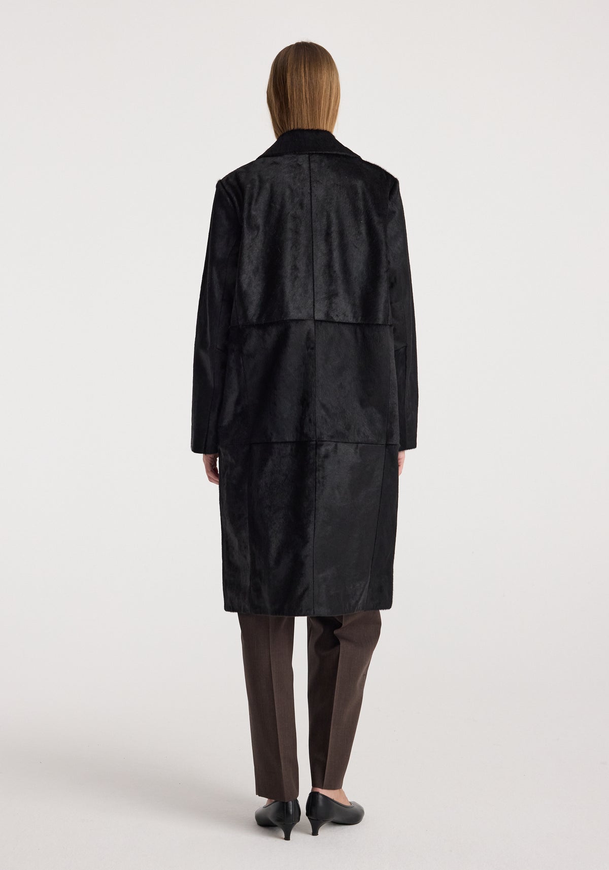Woman wearing a long black textured Black ponyhair coat | black, shown from the back, paired with dark pants and black heeled shoes against a plain background.