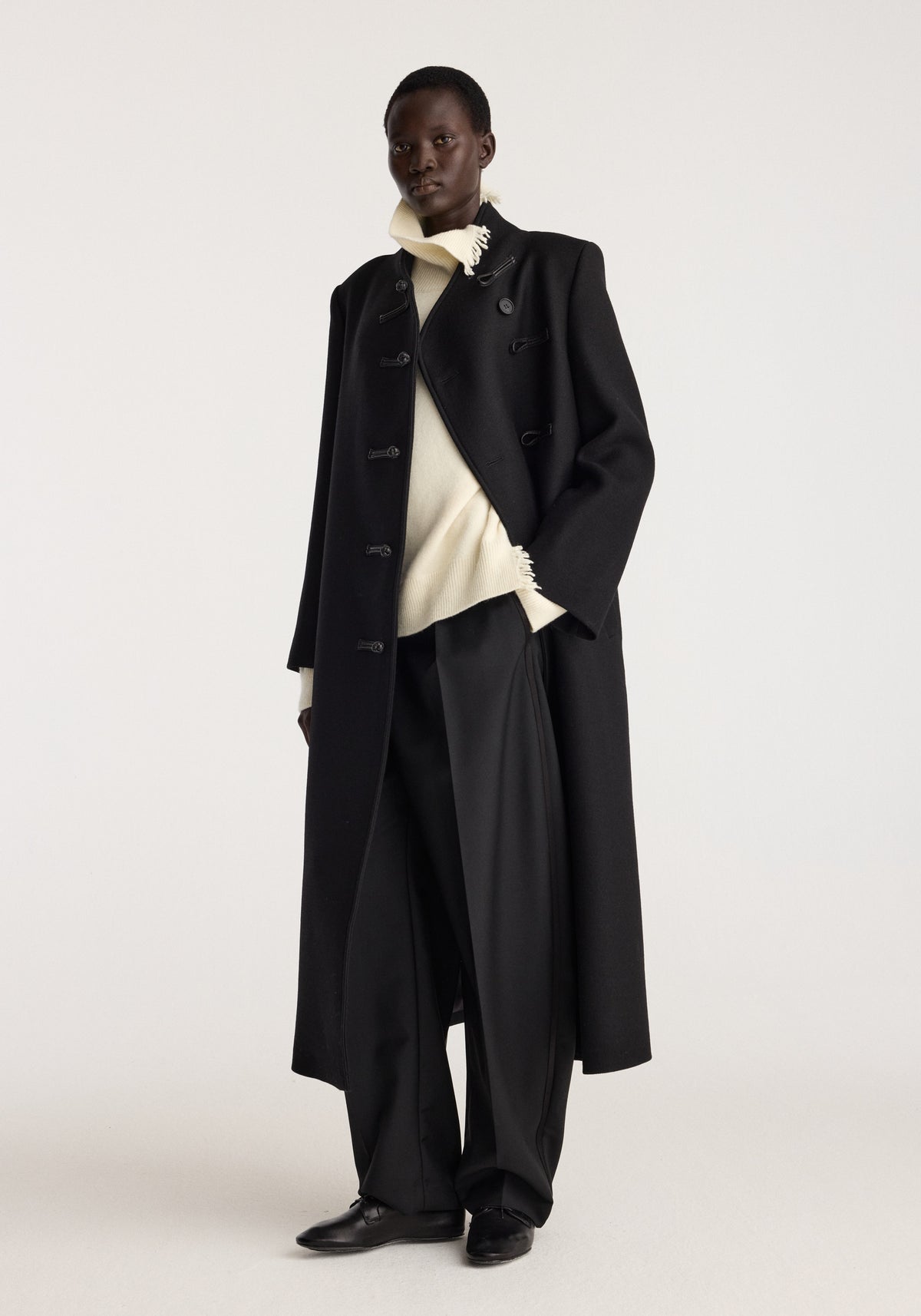 Pankou closure wool coat | black worn by a model over a cream sweater with wide black trousers and black loafers in a studio setting