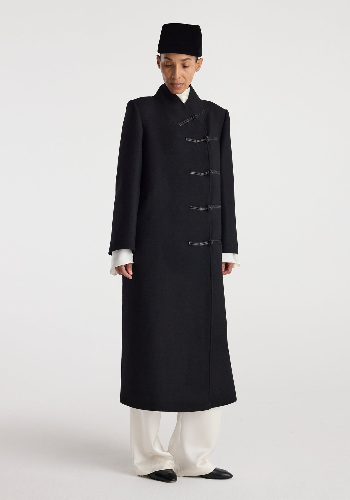 Model wearing a long black wool coat with traditional pankou closures, paired with white pants and black shoes, standing against a plain light background in the Pankou closure wool coat | black.