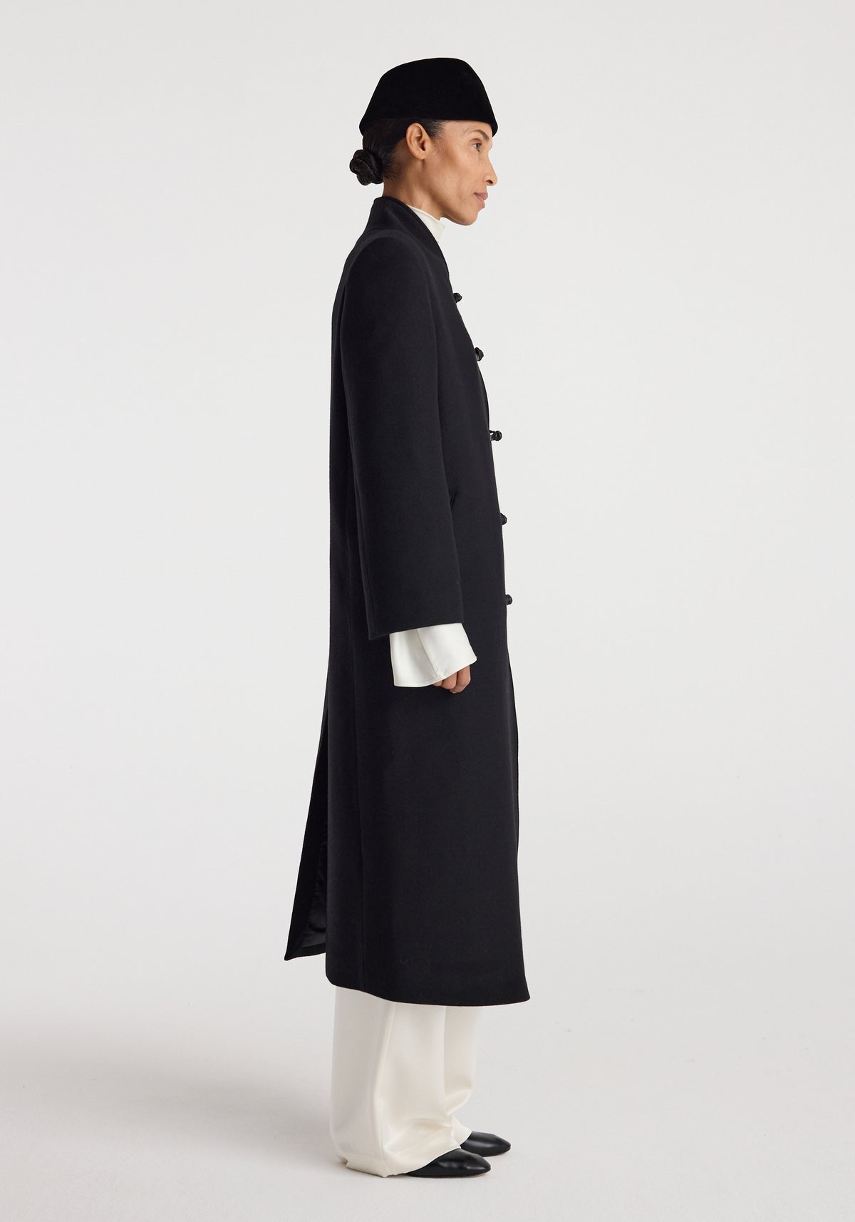 Side view of a person wearing a long black Pankou closure wool coat | black with wide sleeves over white pants and black shoes, paired with a black hat.