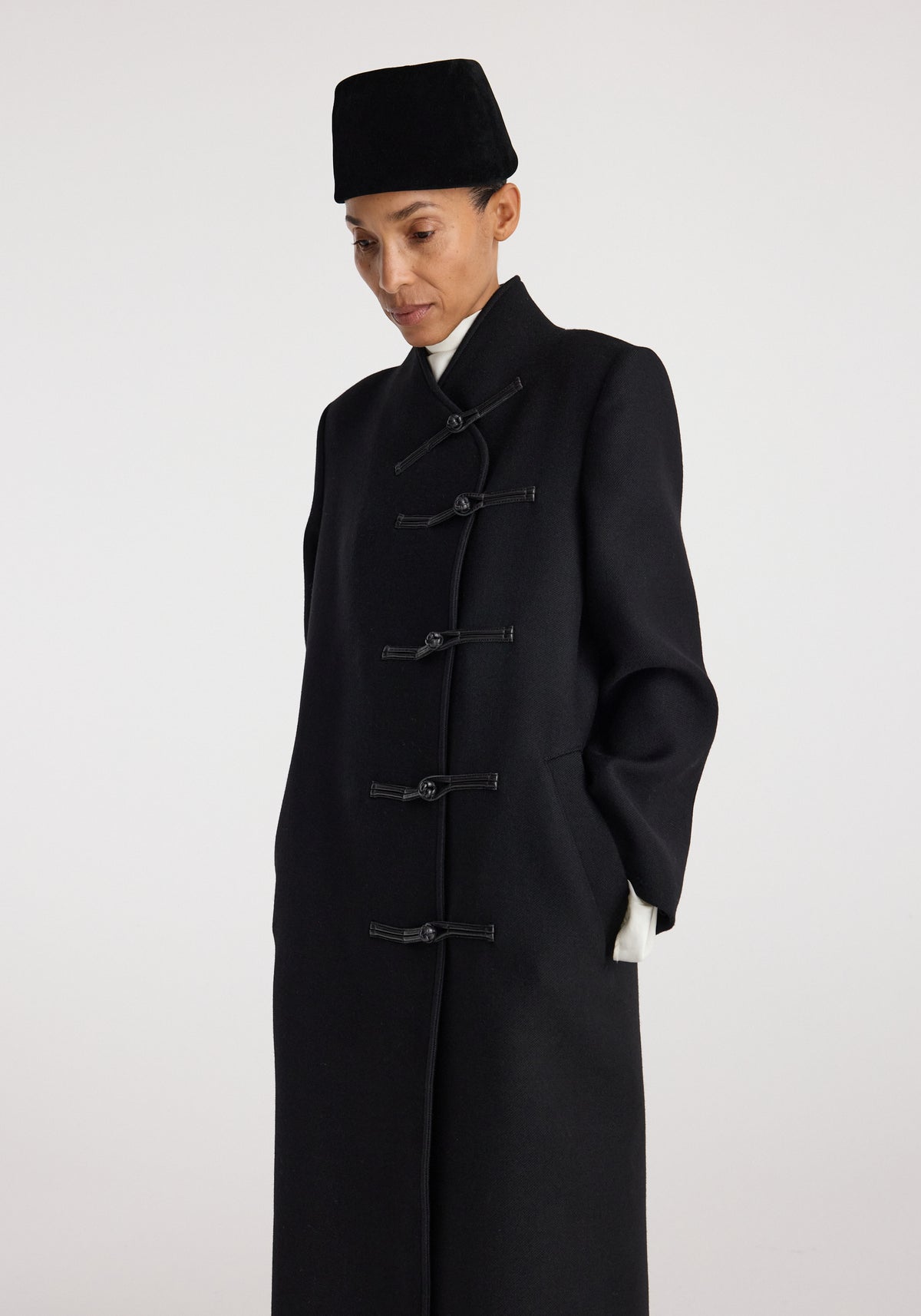 Model wearing a black Pankou closure wool coat | black with traditional knot buttons and a high collar, styled with a black hat against a plain background.