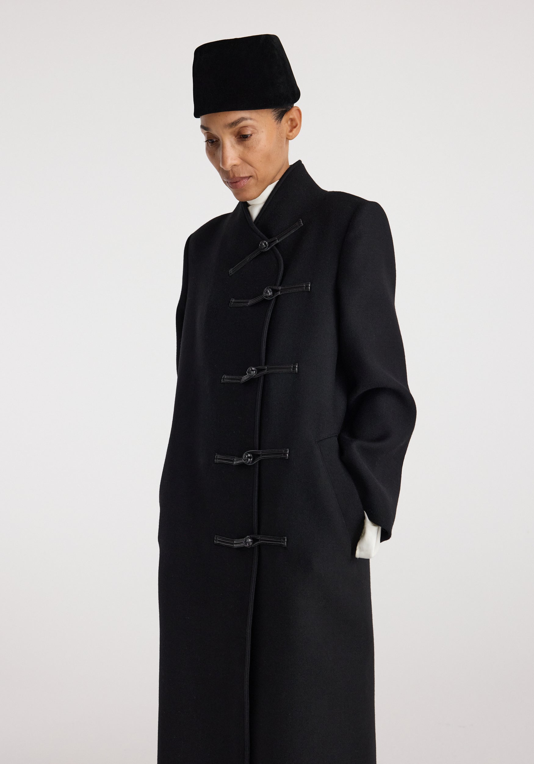 Model wearing a black Pankou closure wool coat | black with traditional knot buttons and a high collar, styled with a black hat against a plain background.