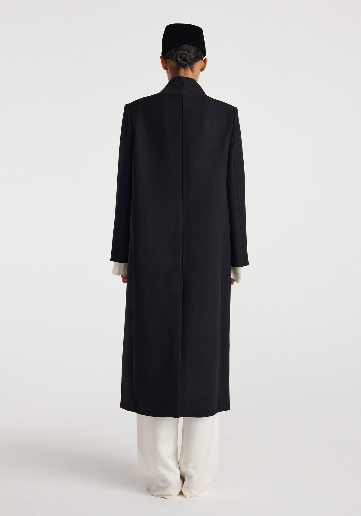 Rear view of a model wearing a long black Pankou closure wool coat | black with a clean, minimalist design, paired with white pants and a black hat against a plain background.