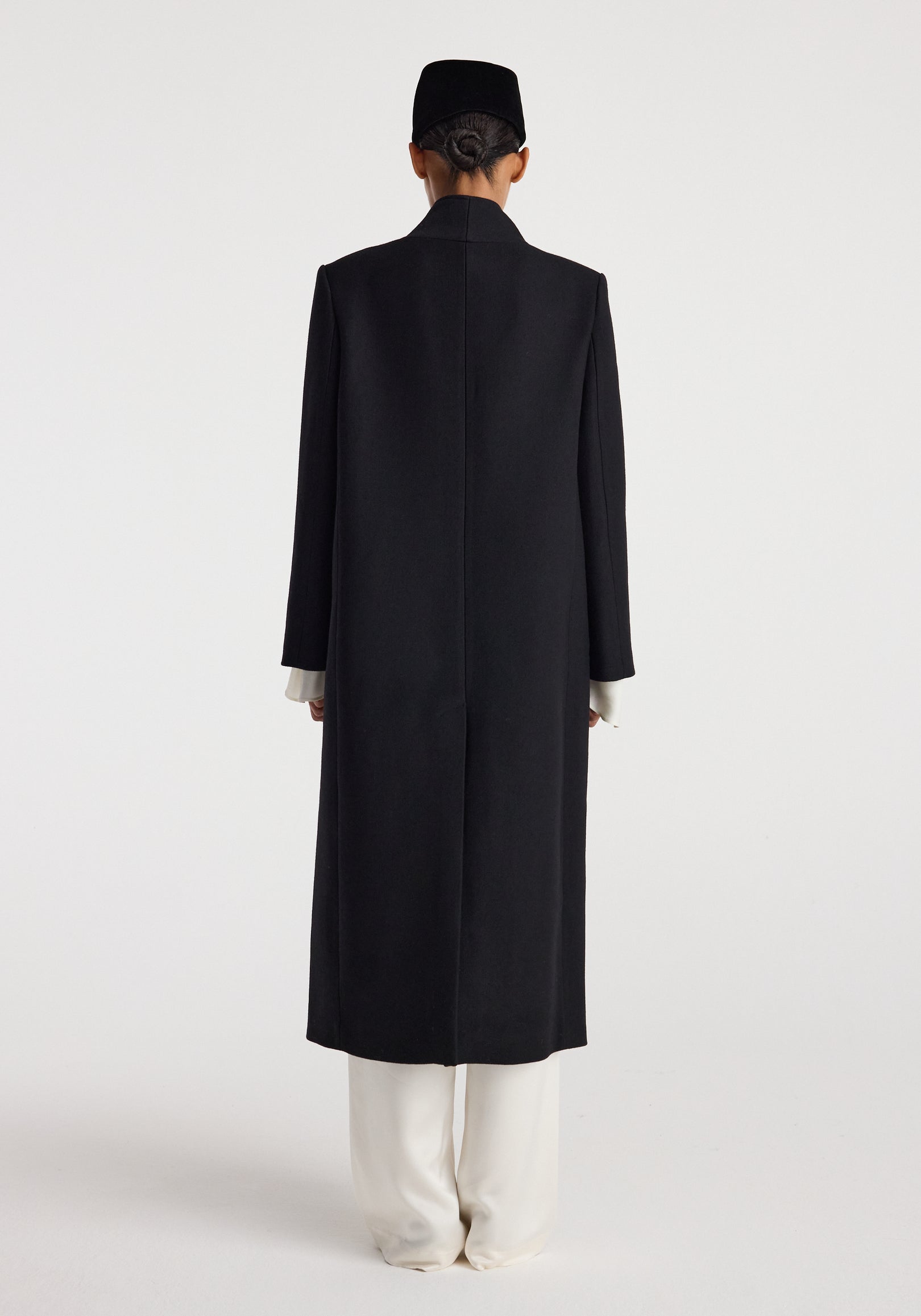 Rear view of a model wearing a long black Pankou closure wool coat | black with a clean, minimalist design, paired with white pants and a black hat against a plain background.