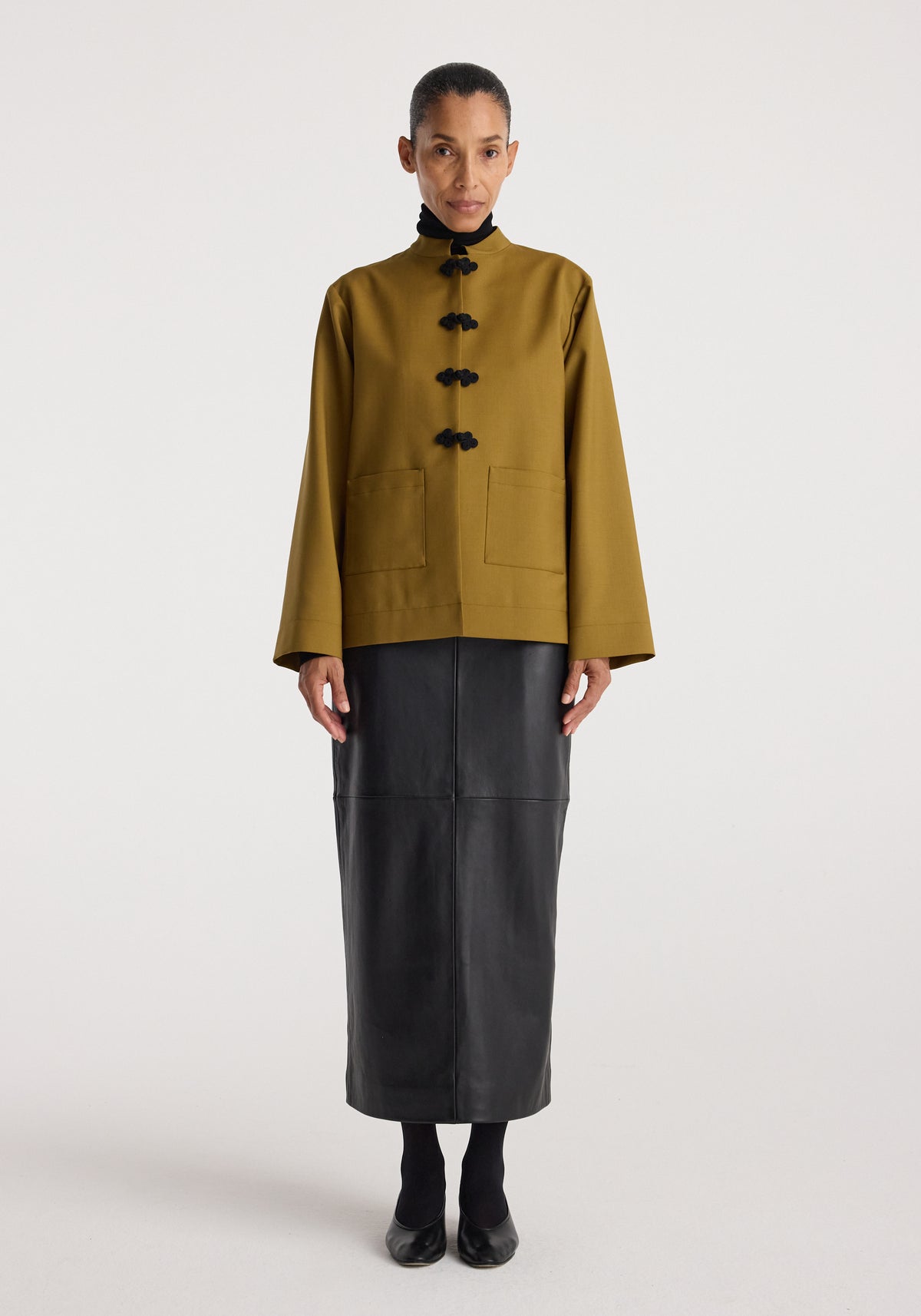 Woman wearing a mustard colored Pankou closure jacket with a high collar and black toggle buttons, paired with a long black skirt and black shoes.