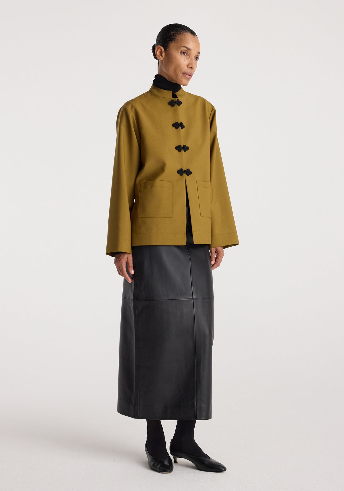 Woman wearing a Pankou closure jacket | mustard with black frog closures, paired with a black turtleneck and a long black leather skirt, standing against a plain background.