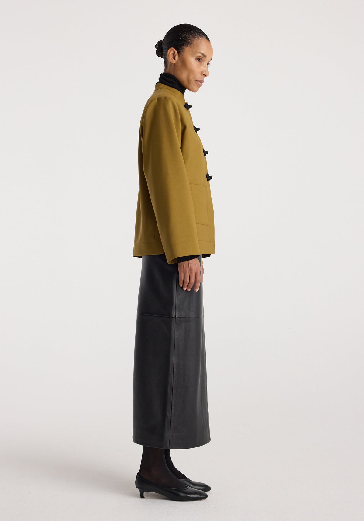 Woman wearing a mustard Pankou closure jacket with black knotted buttons, paired with a long black skirt and black heels, standing in profile against a plain background.