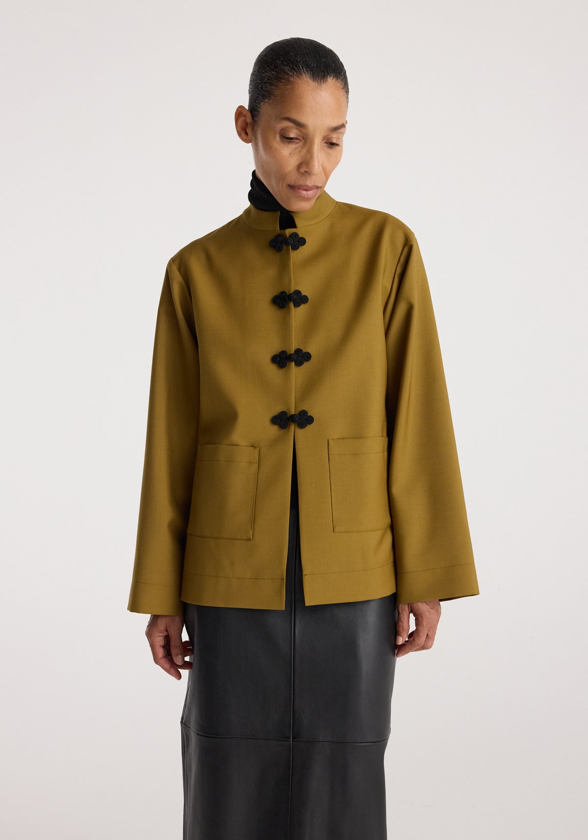 Woman wearing a mustard-colored Pankou closure jacket with black traditional frog button details and front patch pockets, styled with a black skirt.