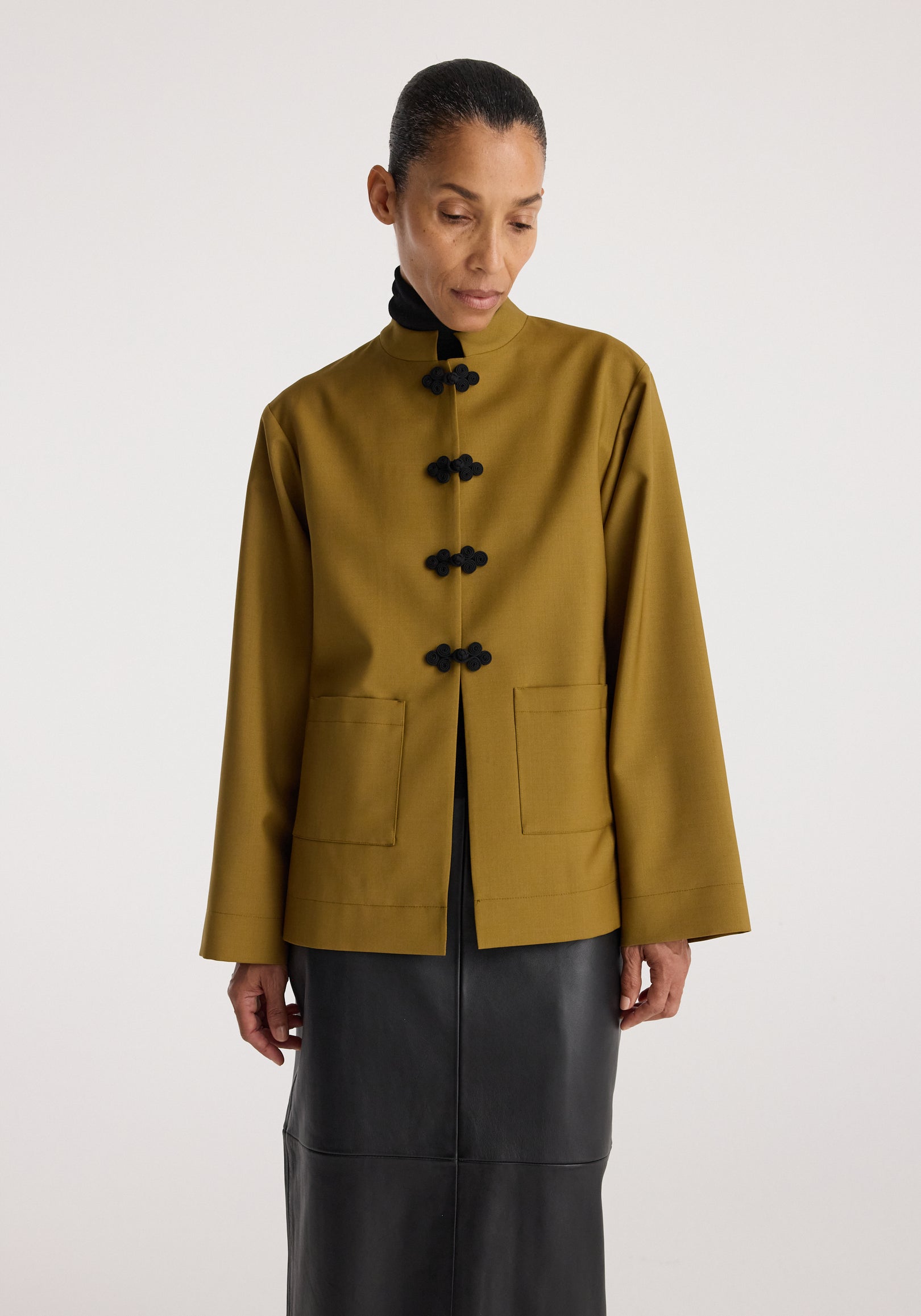 Woman wearing a mustard-colored Pankou closure jacket with black traditional frog button details and front patch pockets, styled with a black skirt.
