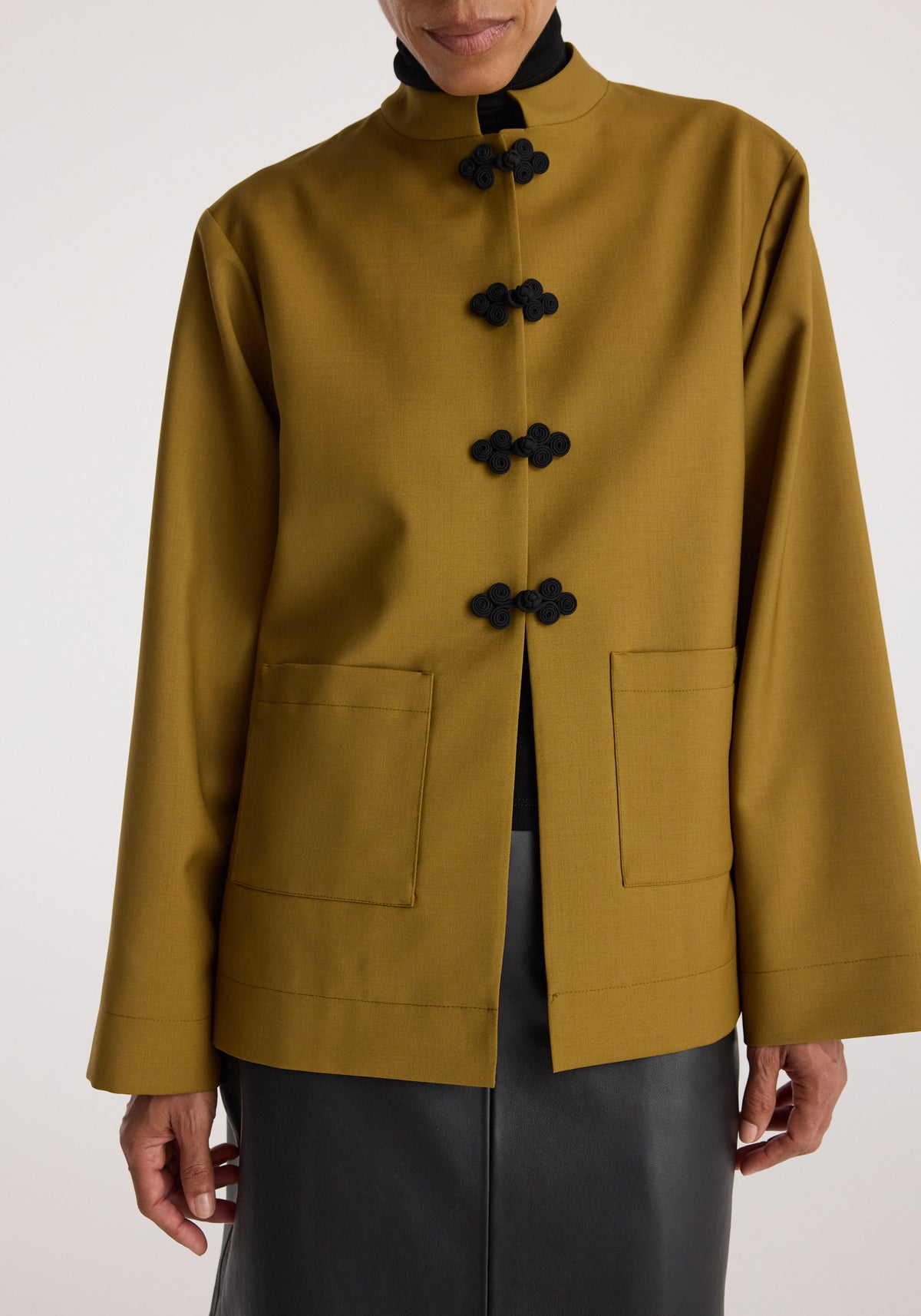 Close-up of a person wearing a mustard Pankou closure jacket with black traditional knot buttons, square front pockets, and a stand collar over a black turtleneck.
