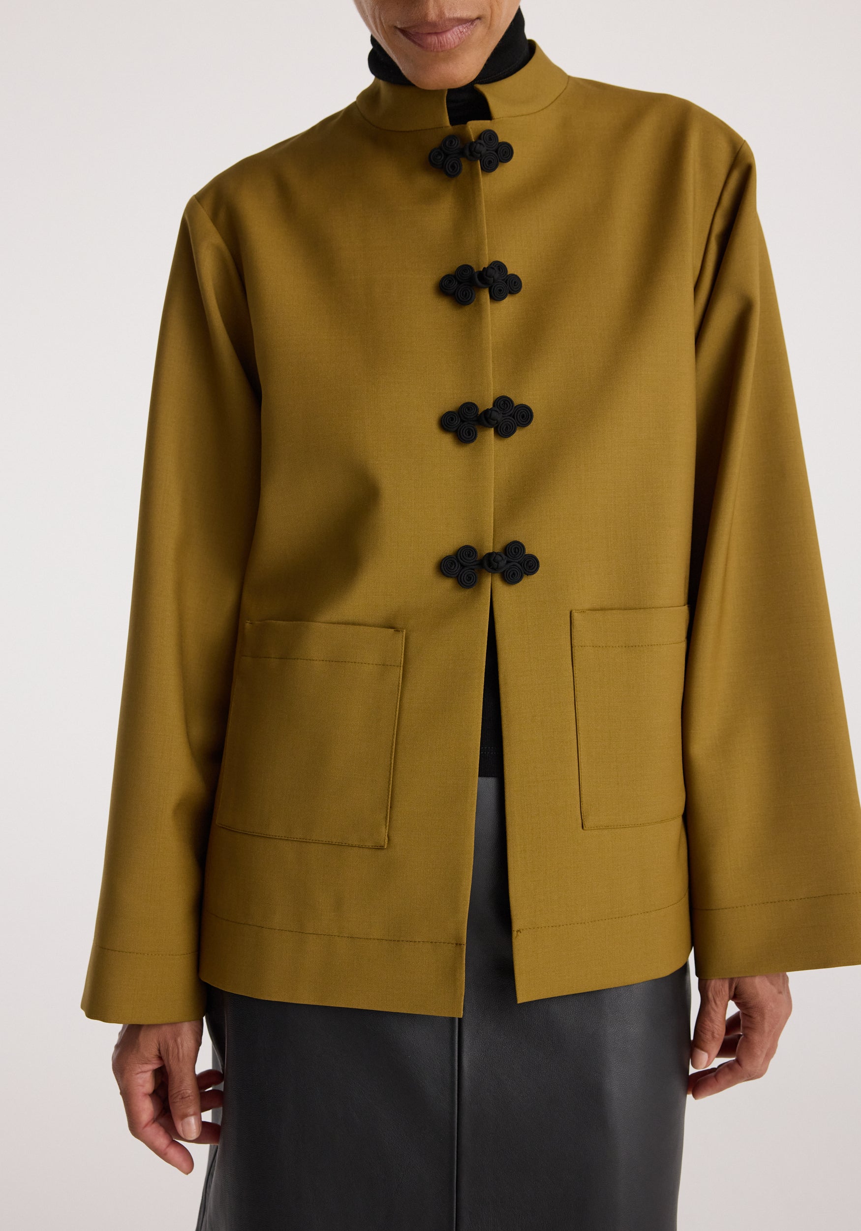 Close-up of a person wearing a mustard Pankou closure jacket with black traditional knot buttons, square front pockets, and a stand collar over a black turtleneck.