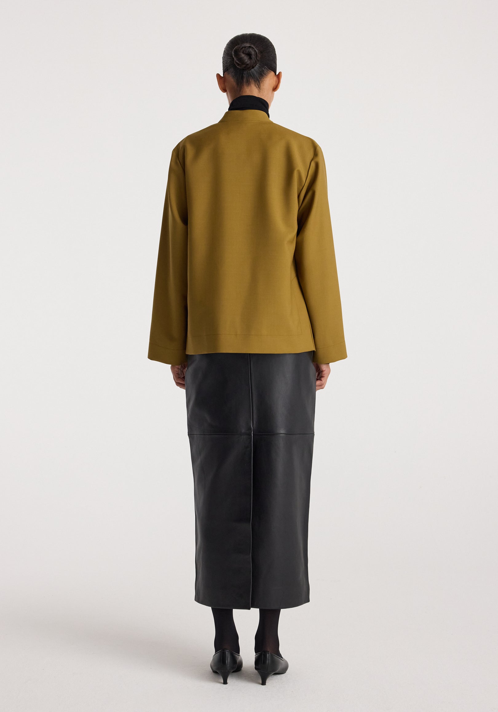 Rear view of a person wearing a mustard colored Pankou closure jacket paired with a black leather skirt and black heels against a plain background.
