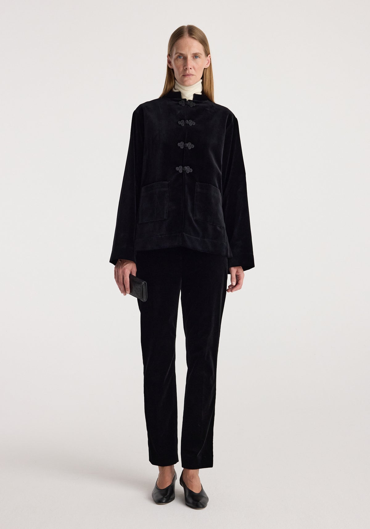 Woman wearing a black Pankou closure velvet jacket | black with matching black velvet pants, holding a black clutch and standing against a plain background.