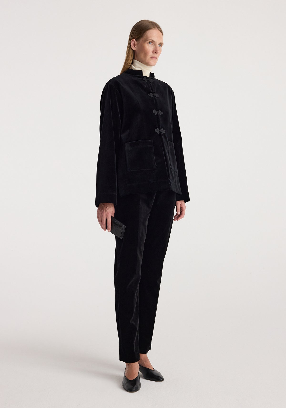 Woman wearing a black velvet jacket with pankou closure over a light turtleneck, paired with black velvet pants and black shoes, standing against a white background, showcasing the Pankou closure velvet jacket | black.