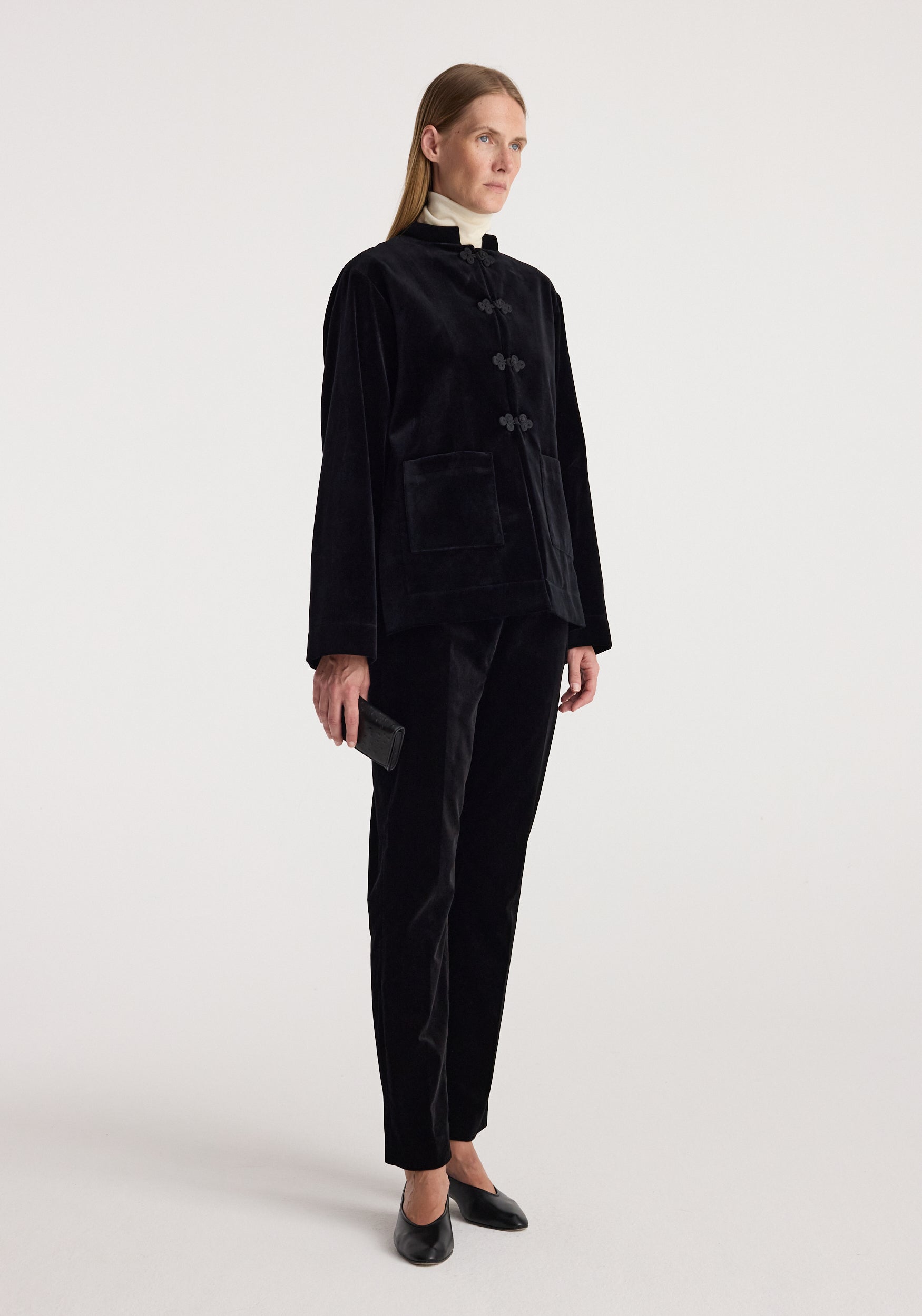 Woman wearing a black velvet jacket with pankou closure over a light turtleneck, paired with black velvet pants and black shoes, standing against a white background, showcasing the Pankou closure velvet jacket | black.