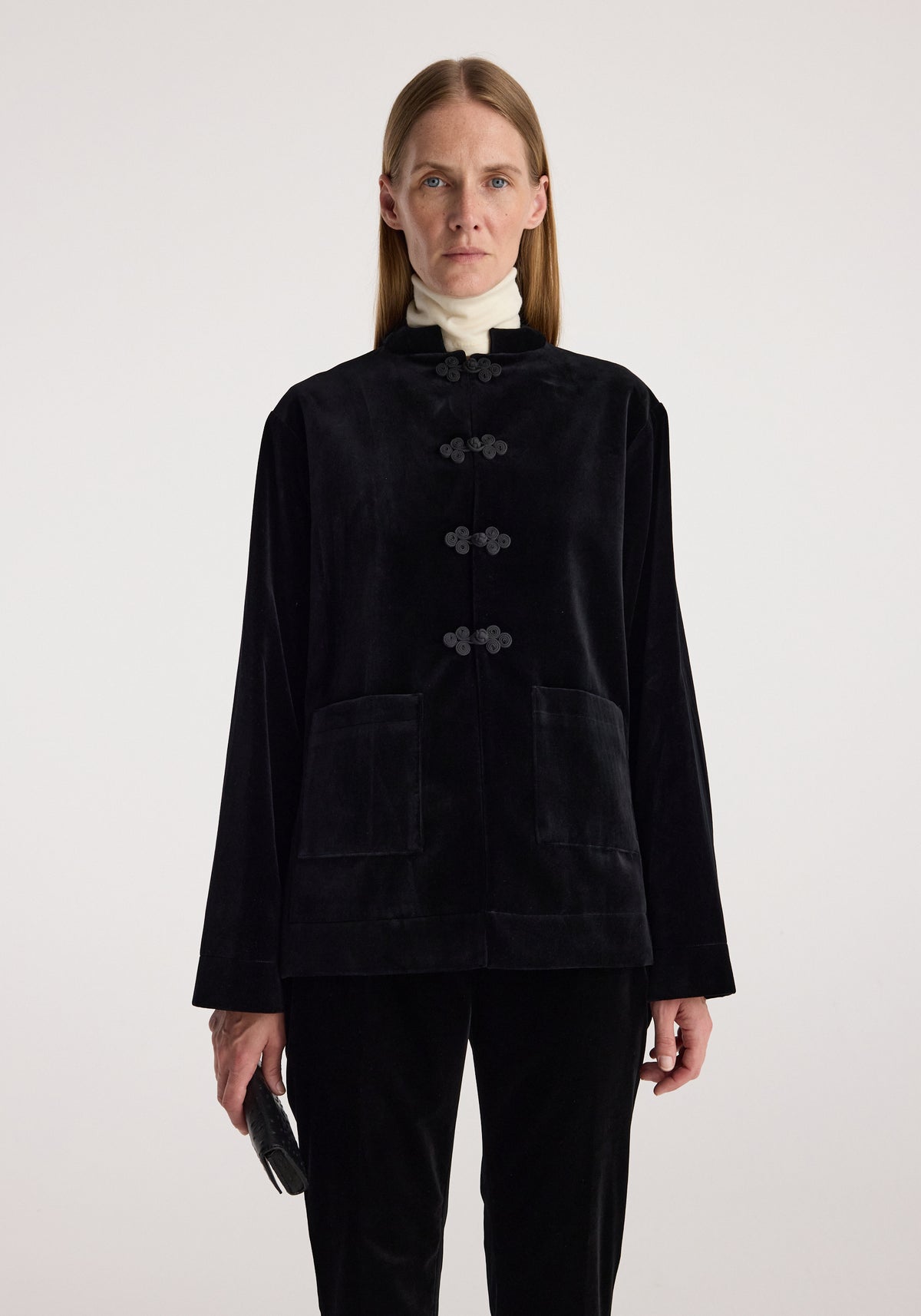 Woman wearing a black Pankou closure velvet jacket | black with ornamental knot buttons and large front pockets, paired with black pants and a white turtleneck.