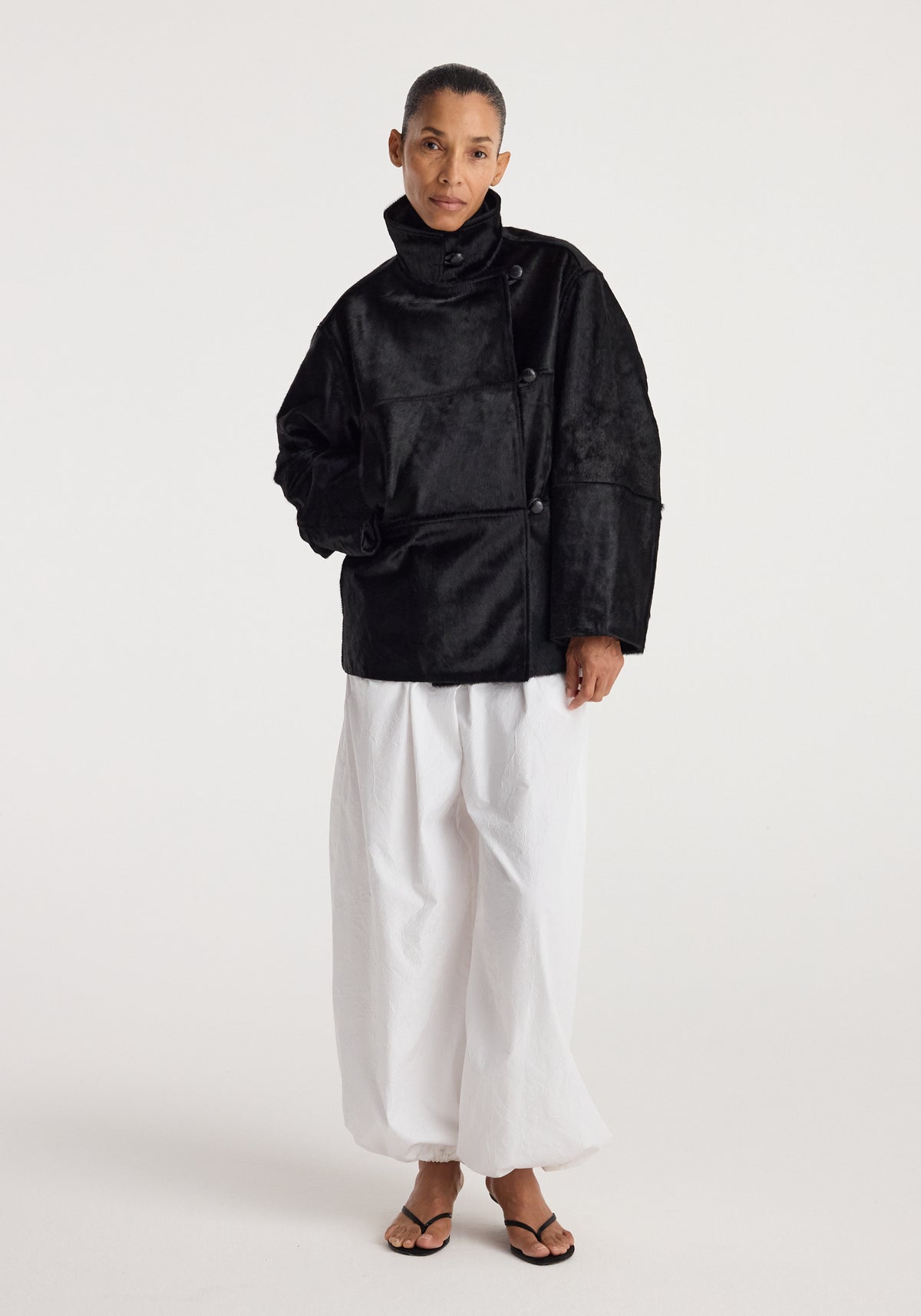 Woman wearing a black ponyhair jacket | black with a high collar and button details paired with loose white pants and black sandals against a plain background