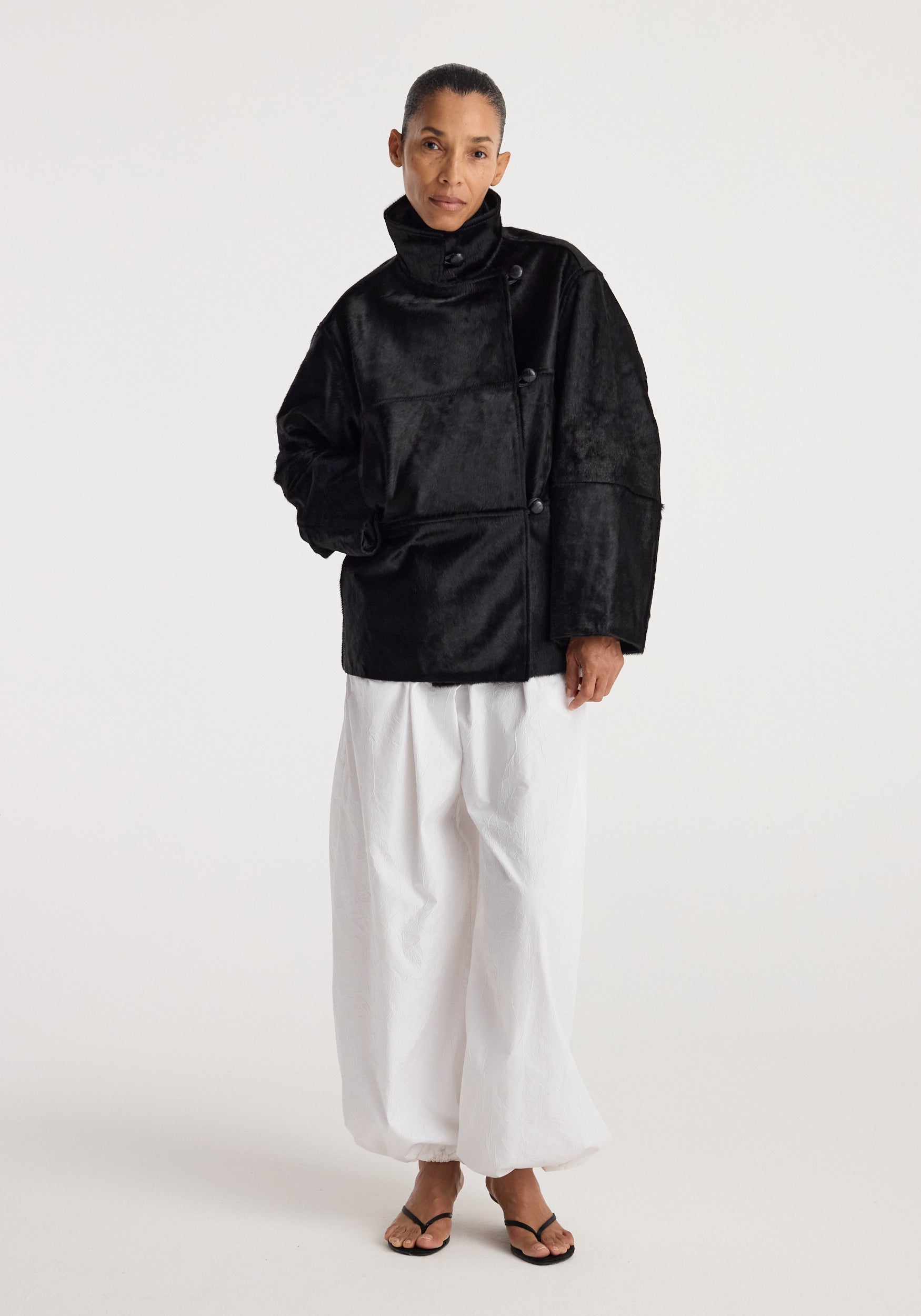 Woman wearing a black ponyhair jacket | black with a high collar and button details paired with loose white pants and black sandals against a plain background