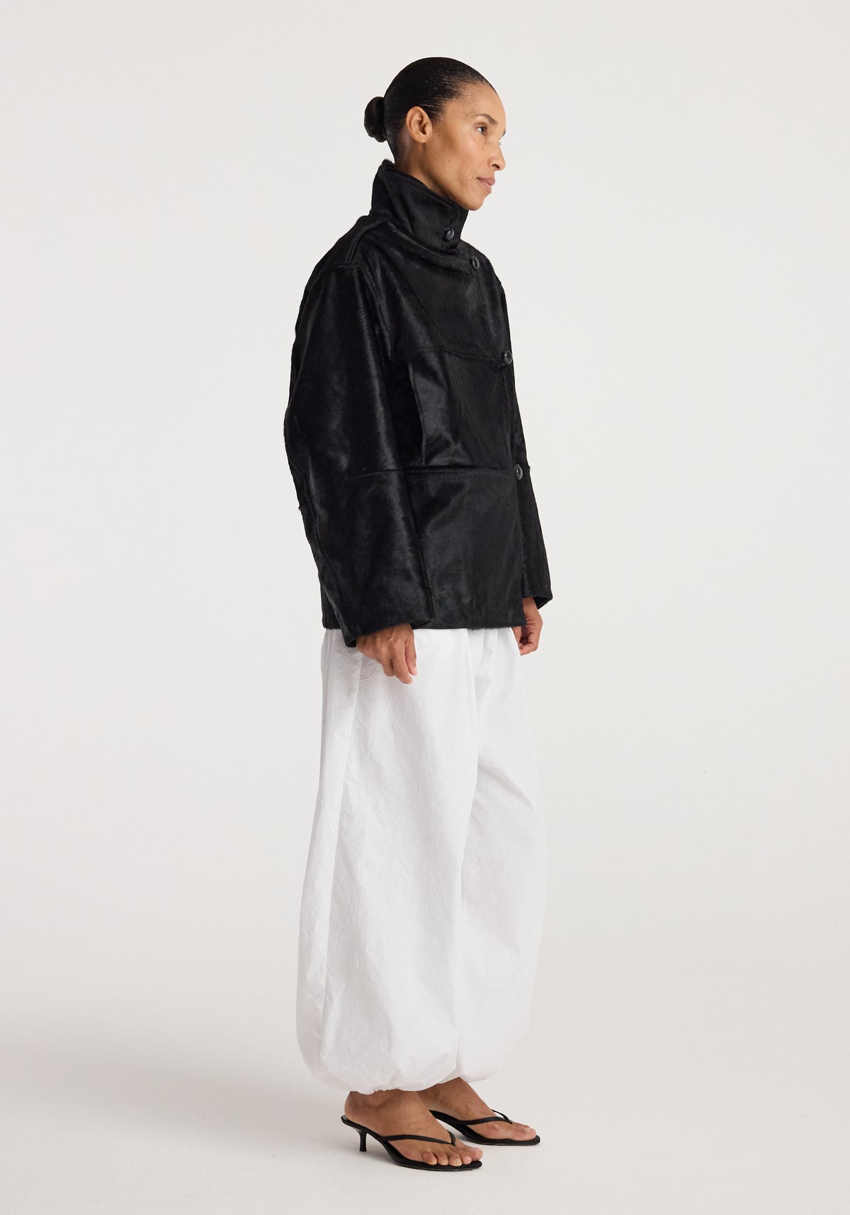 Model wearing a Black ponyhair jacket | black with high collar and button closure, paired with loose white pants and black heeled sandals against a plain background.