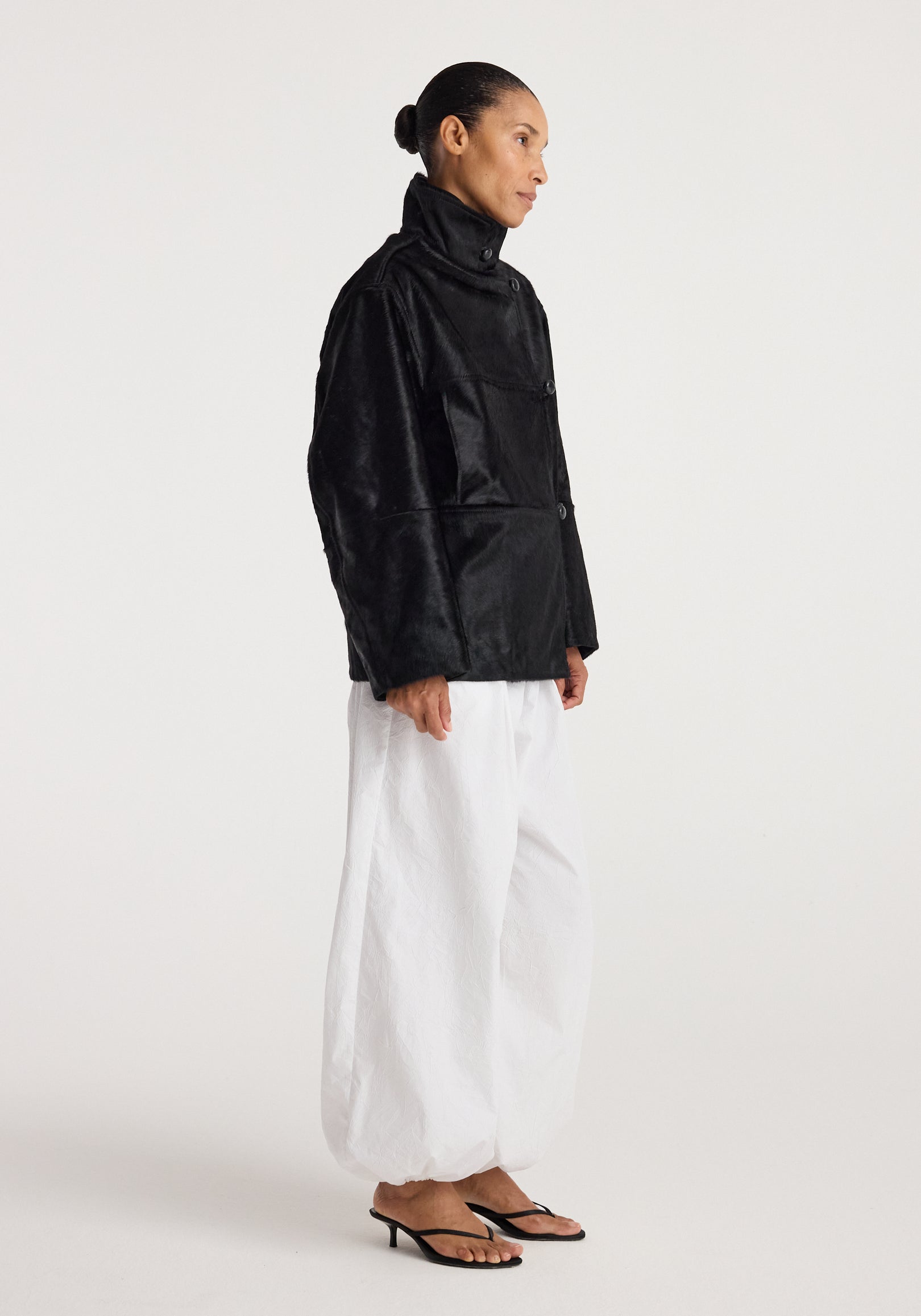 Model wearing a Black ponyhair jacket | black with high collar and button closure, paired with loose white pants and black heeled sandals against a plain background.