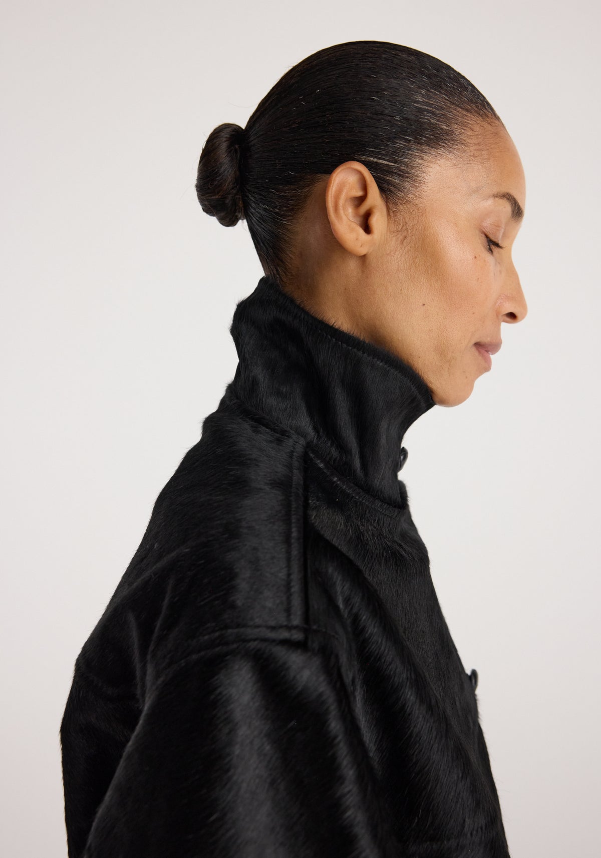 Profile view of a woman wearing a high-collar Black ponyhair jacket | black with sleek hair pulled back into a bun against a plain background.