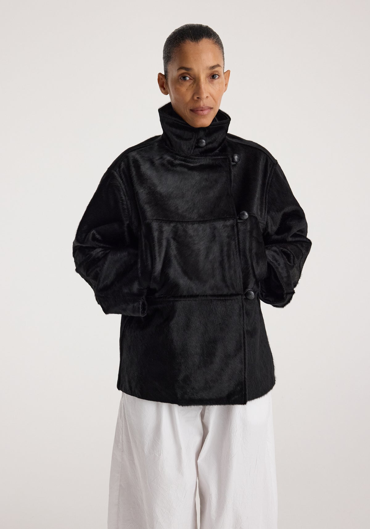 Woman wearing a black ponyhair jacket | black with a high collar and button details, styled with loose white pants against a plain background.