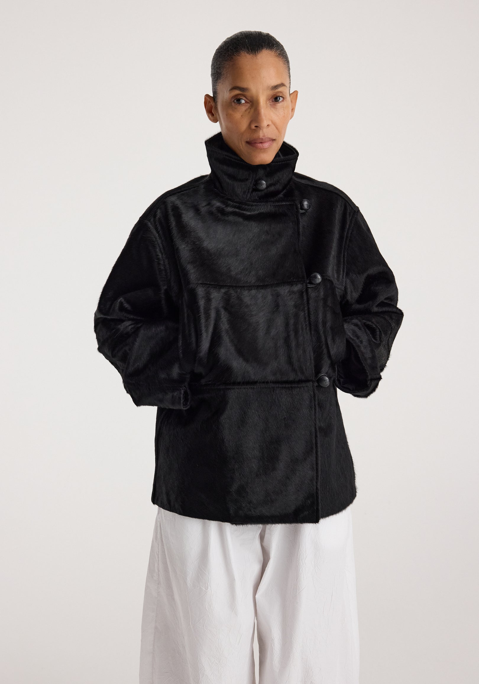 Woman wearing a black ponyhair jacket | black with a high collar and button details, styled with loose white pants against a plain background.