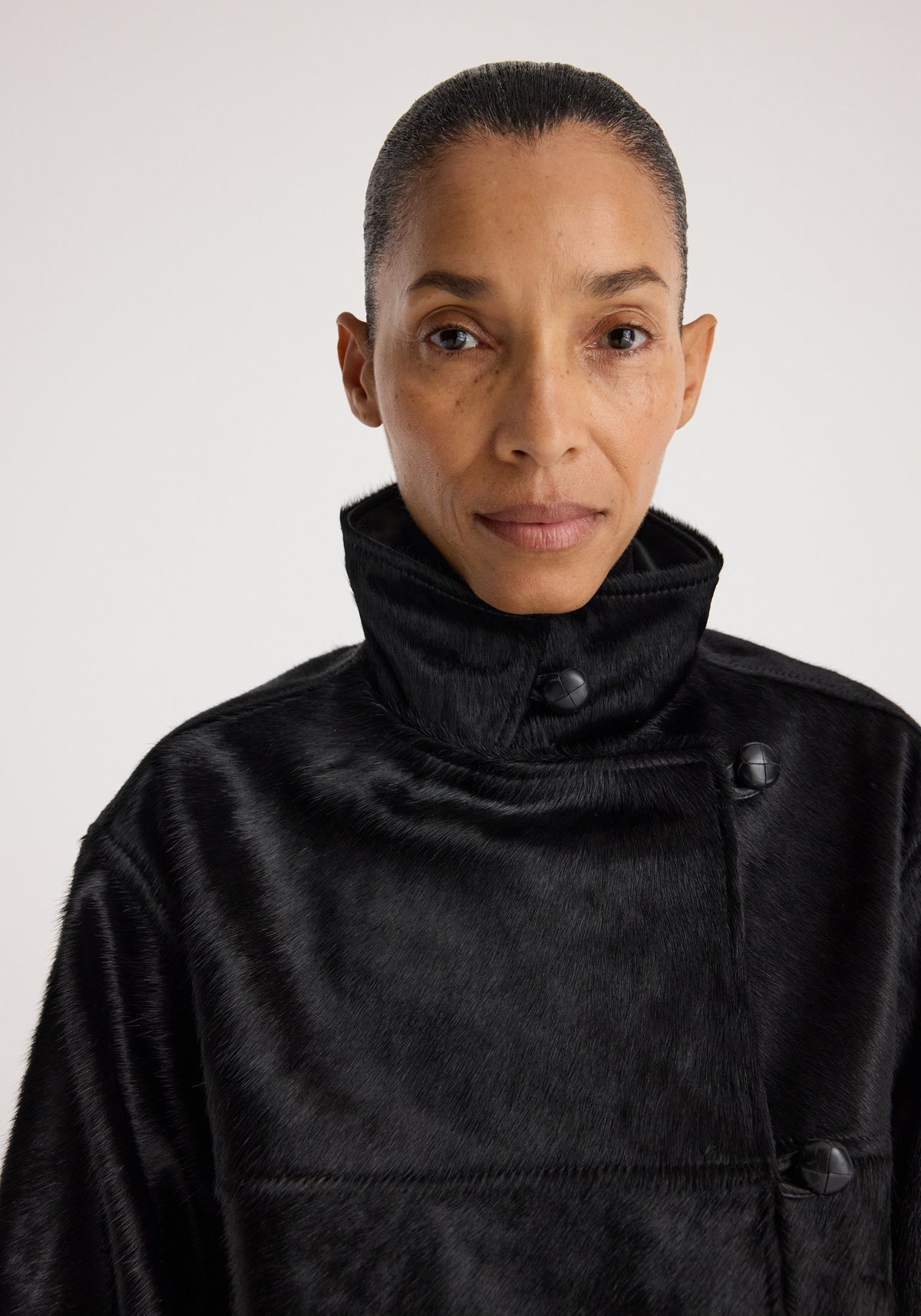 Close-up portrait of a person wearing a Black ponyhair jacket | black with a high collar and visible buttons against a plain background.