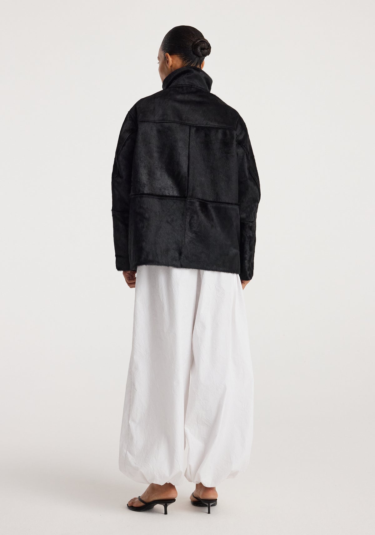 Back view of a person wearing a Black ponyhair jacket | black with a high collar paired with loose white pants and black heeled sandals against a plain background.