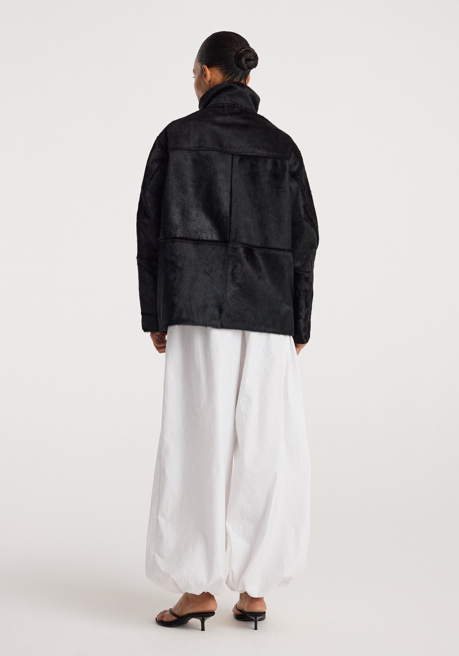 Back view of a person wearing a Black ponyhair jacket | black with a high collar paired with loose white pants and black heeled sandals against a plain background.