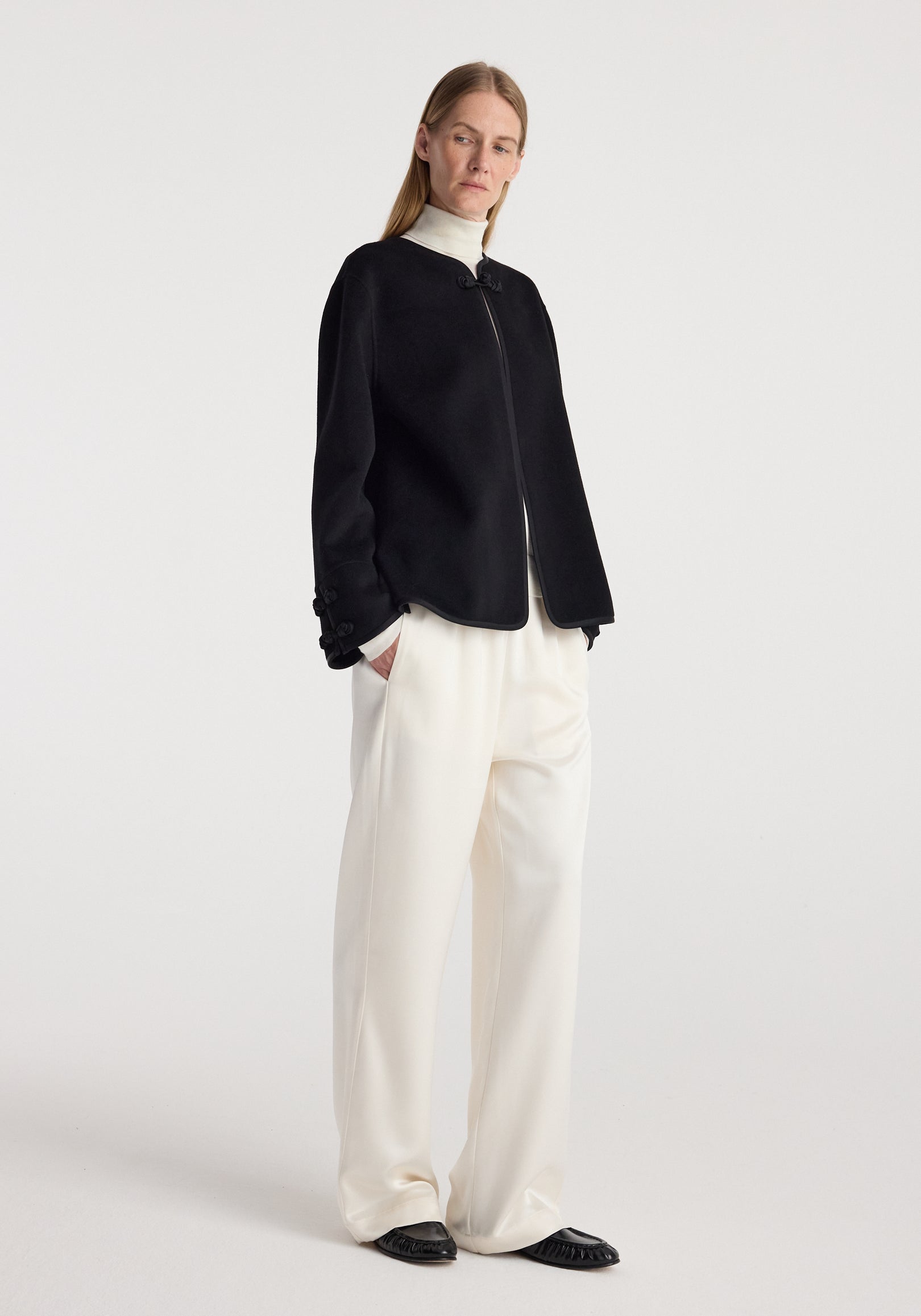 Woman wearing a black Double-faced pankou jacket | black over a white turtleneck and cream wide-leg pants, standing against a plain white background with hands in pockets.