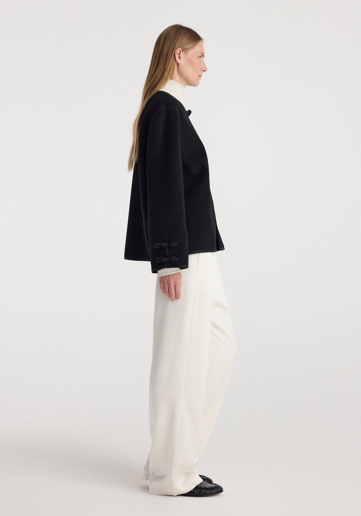 Side profile of a woman wearing a black Double-faced pankou jacket | black with traditional pankou button details paired with white wide-leg pants and black shoes against a plain background.