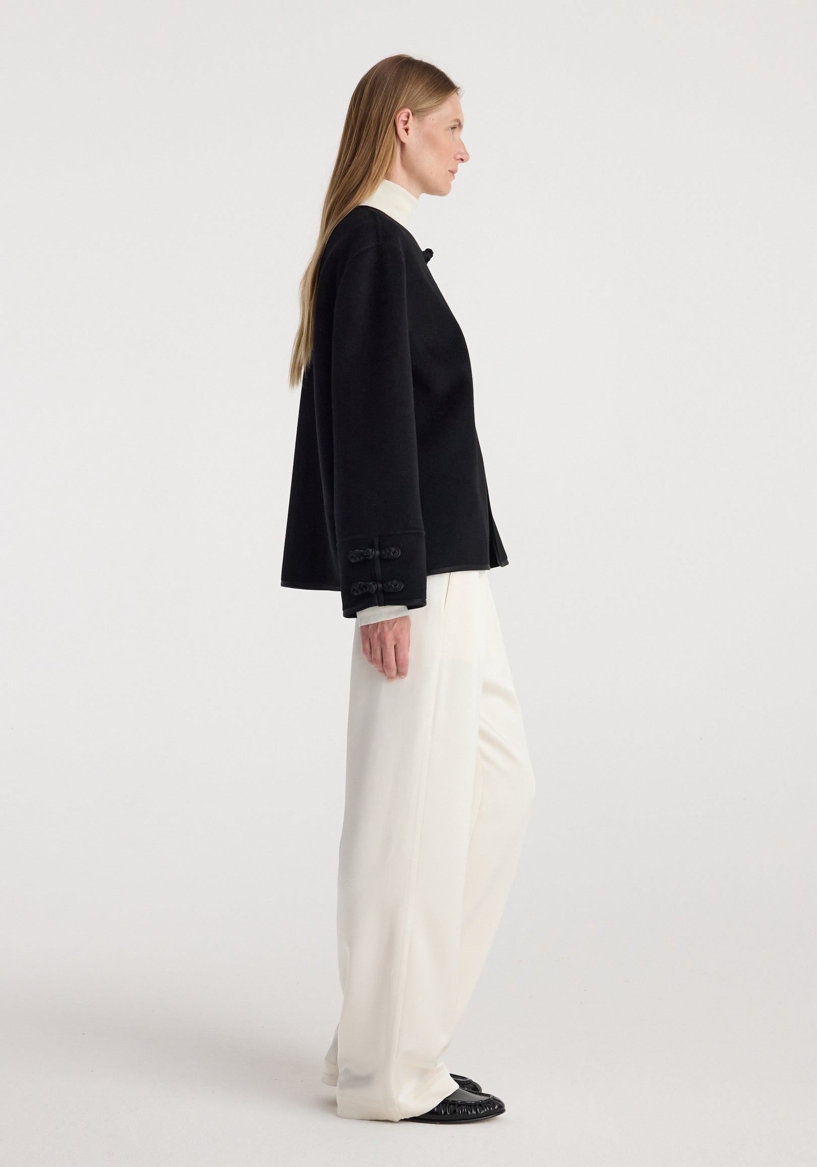 Side profile of a woman wearing a black Double-faced pankou jacket | black with traditional pankou button details paired with white wide-leg pants and black shoes against a plain background.