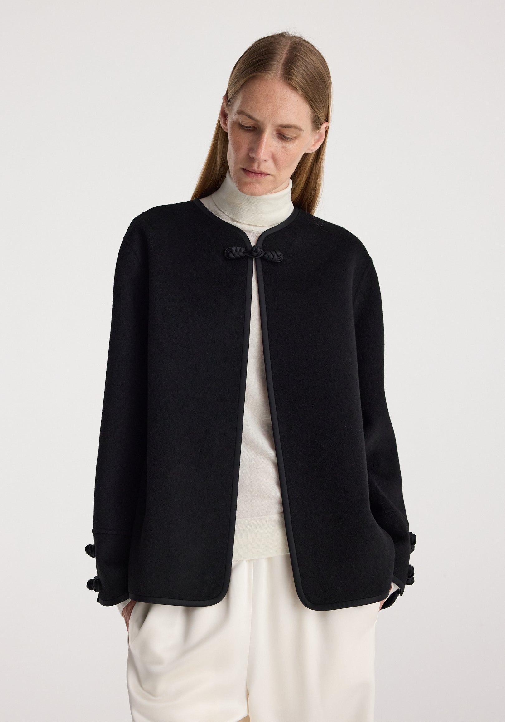 Woman wearing a minimalist black Double-faced pankou jacket | black with traditional pankou knot closures, paired with a white turtleneck and cream pants against a plain background.
