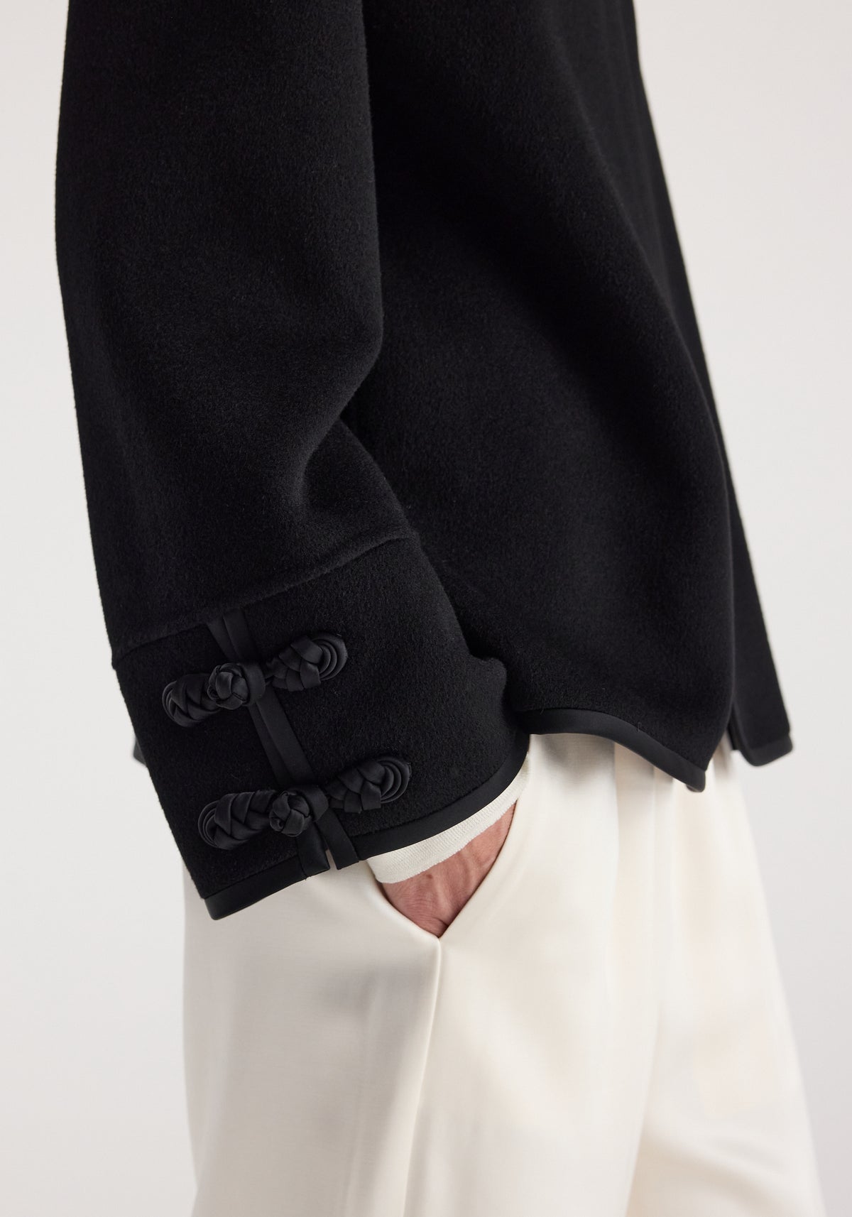 Close-up of a black Double-faced pankou jacket | black sleeve detail with traditional pankou knot buttons, worn with white pants and a hand in the pocket.