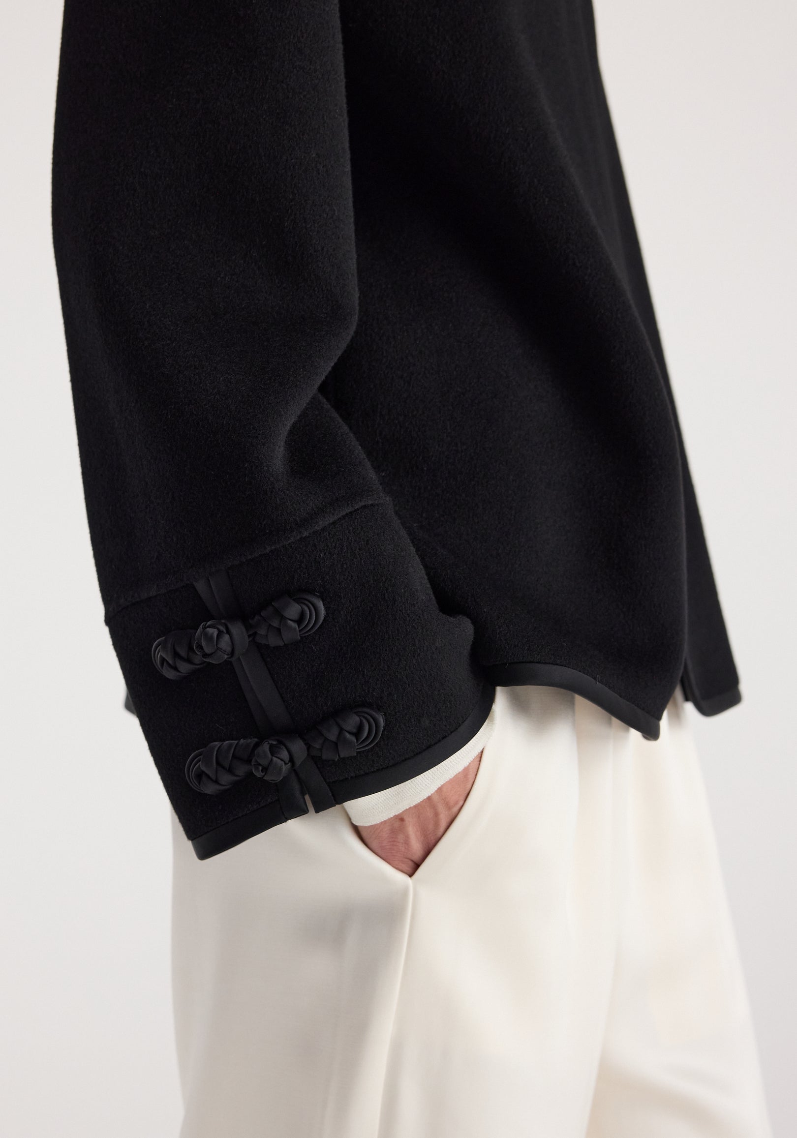 Close-up of a black Double-faced pankou jacket | black sleeve detail with traditional pankou knot buttons, worn with white pants and a hand in the pocket.