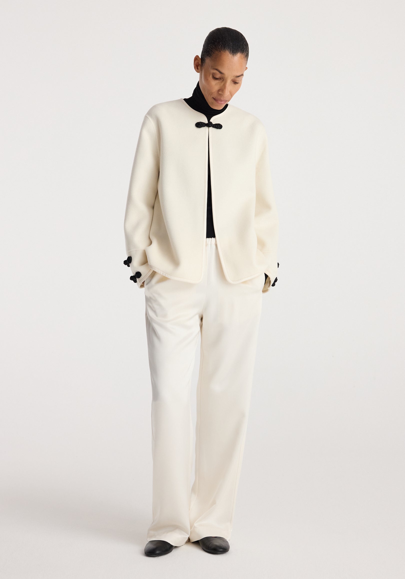 A model wearing a Double-faced pankou jacket | off white with black fastening details, paired with loose off-white pants and black shoes against a plain background.