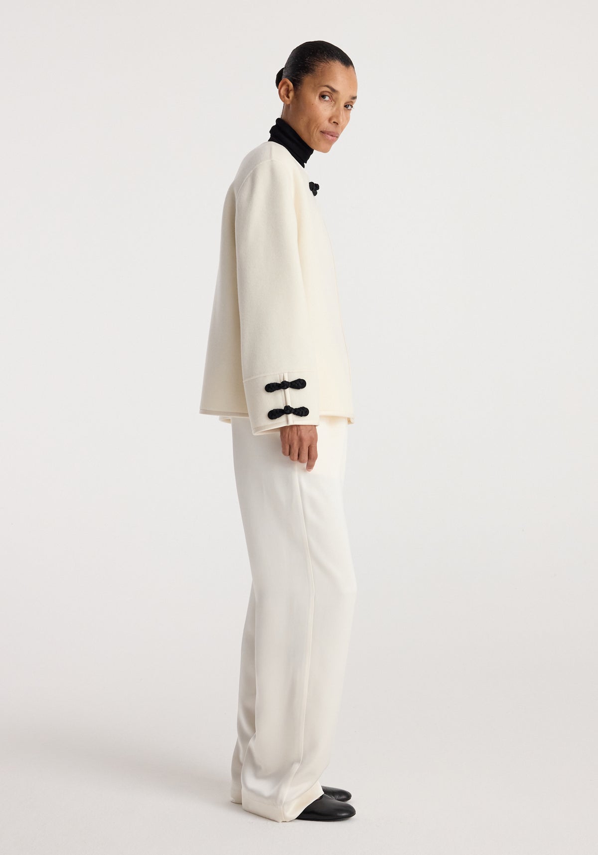 Woman wearing an off white double-faced pankou jacket with black knot closures on the sleeves, paired with white pants and black shoes, standing sideways.