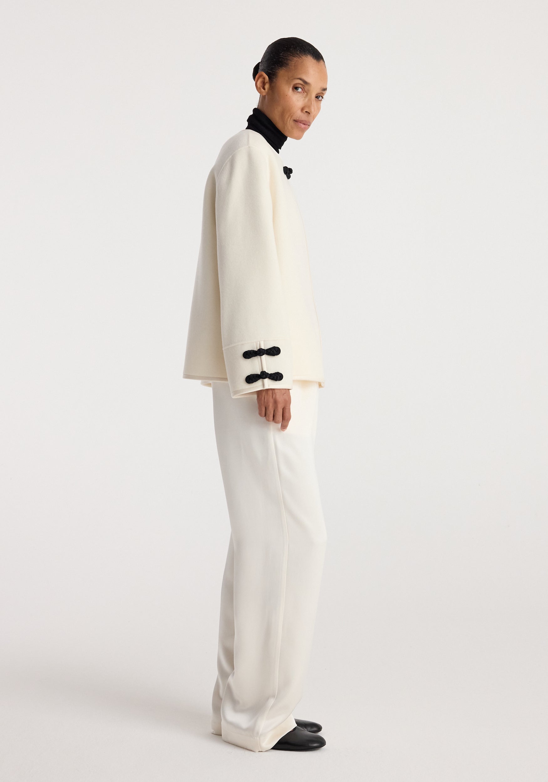Woman wearing an off white double-faced pankou jacket with black knot closures on the sleeves, paired with white pants and black shoes, standing sideways.