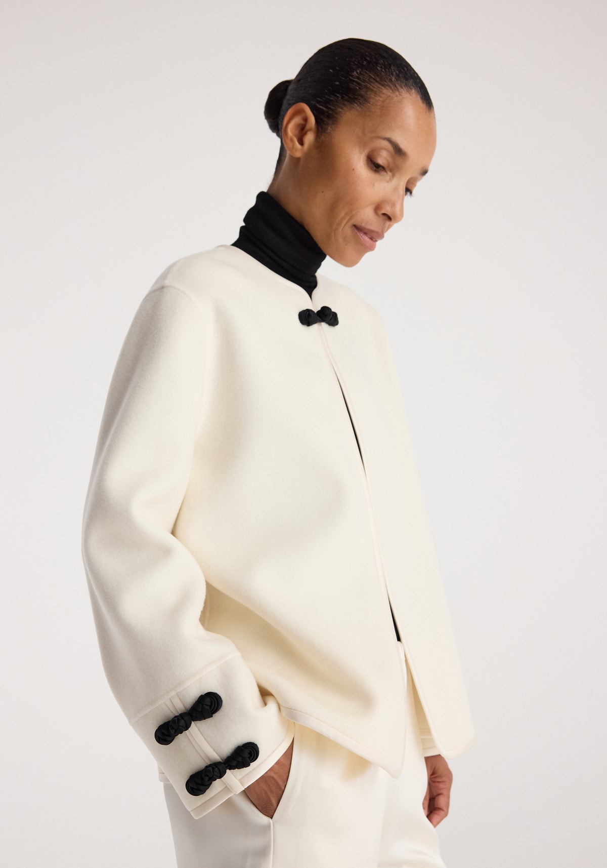 Woman wearing a minimalist off-white Double-faced pankou jacket | off white with black pankou knot buttons paired with a black turtleneck and white pants against a plain background.