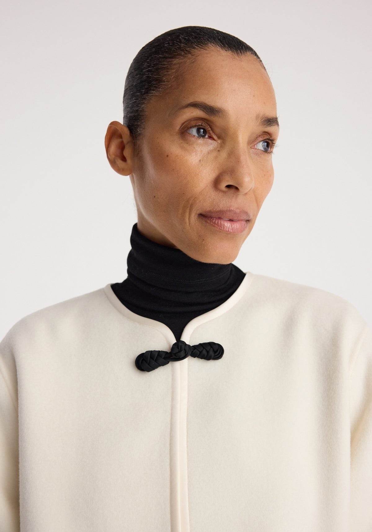 Close-up of a woman wearing a Double-faced pankou jacket | off white with a high collar and a black pankou knot closure, layered over a black turtleneck sweater.