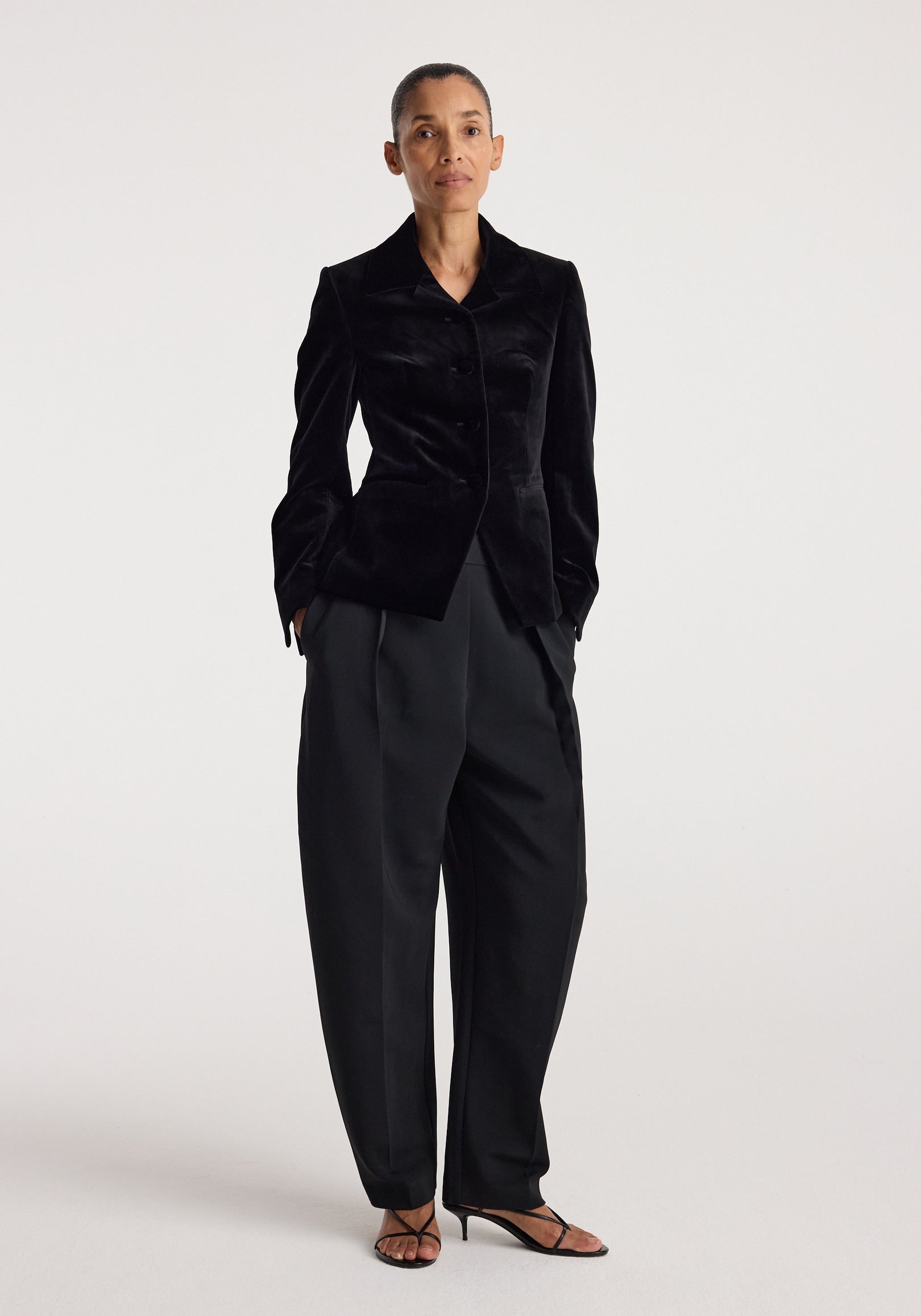 Woman wearing a tailored Classic fitted velvet blazer | black with black wide-leg pants and black strappy heels, standing against a plain white background.