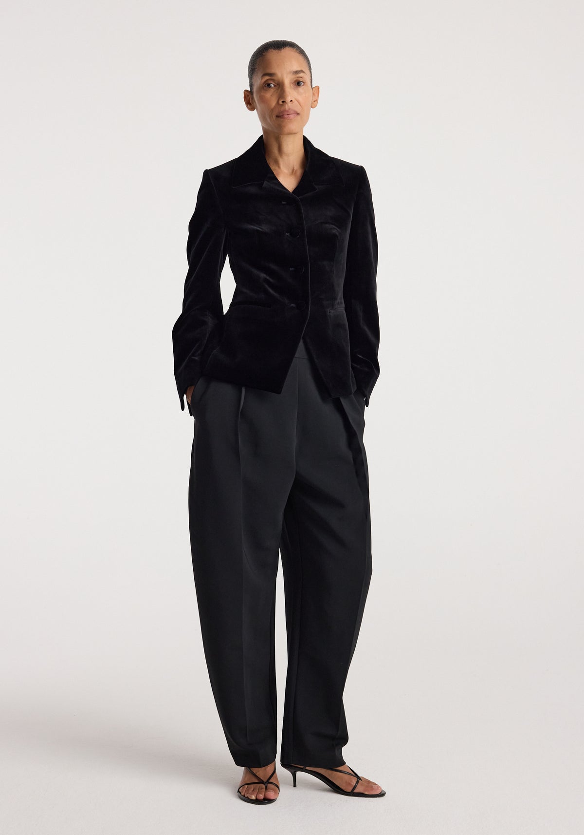 Woman wearing a tailored Classic fitted velvet blazer | black with black wide-leg pants and black strappy heels, standing against a plain white background.
