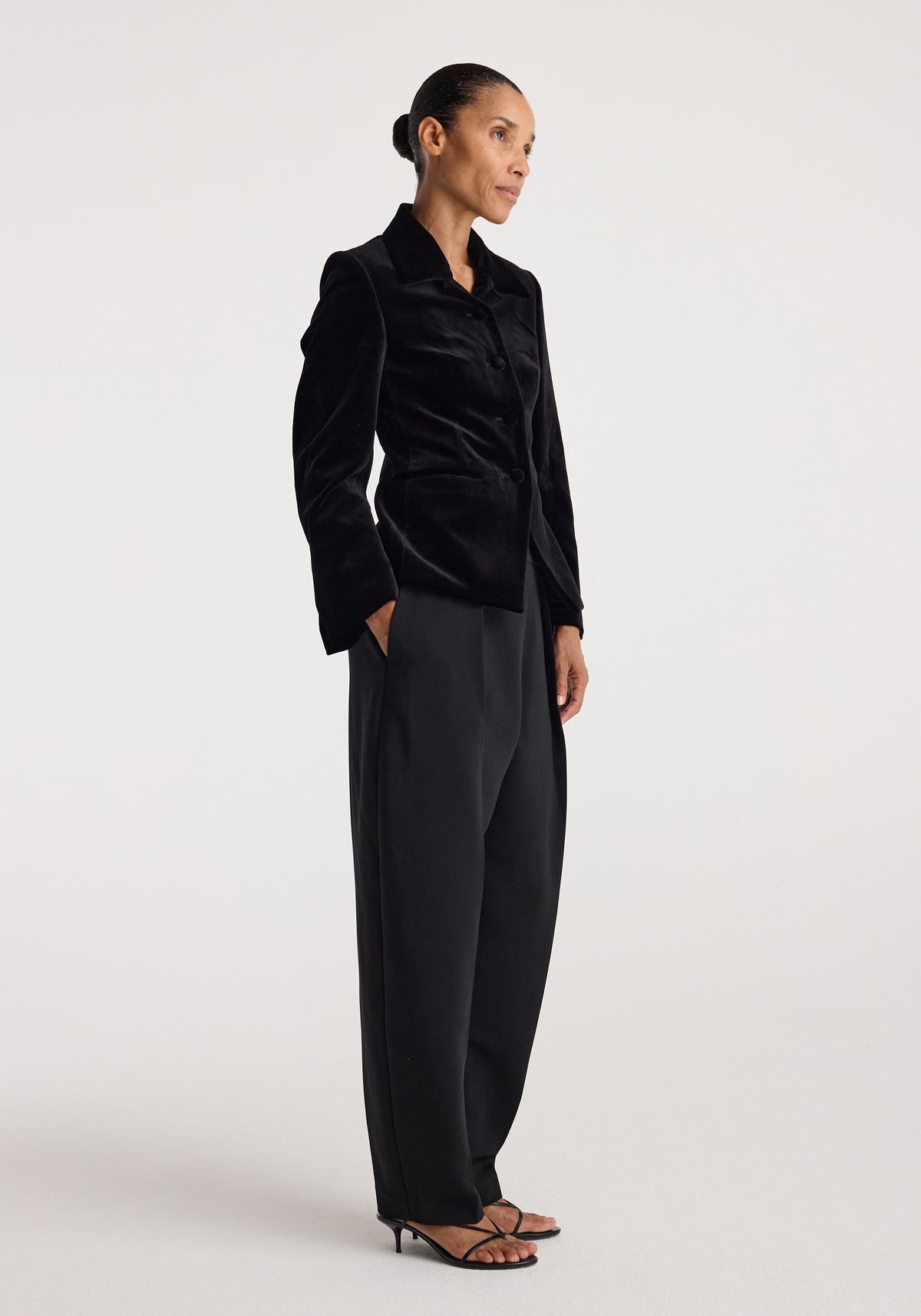Woman wearing a black Classic fitted velvet blazer | black with structured shoulders, button closure, paired with loose black pants and strappy heels against a plain background.