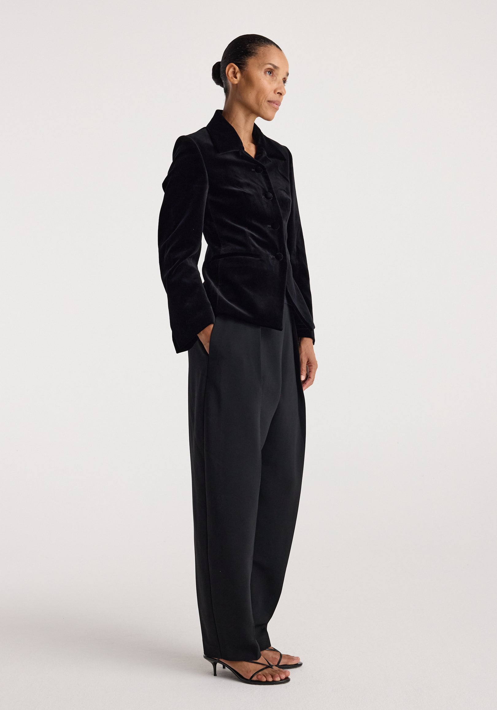 Woman wearing a black Classic fitted velvet blazer | black with structured shoulders, button closure, paired with loose black pants and strappy heels against a plain background.