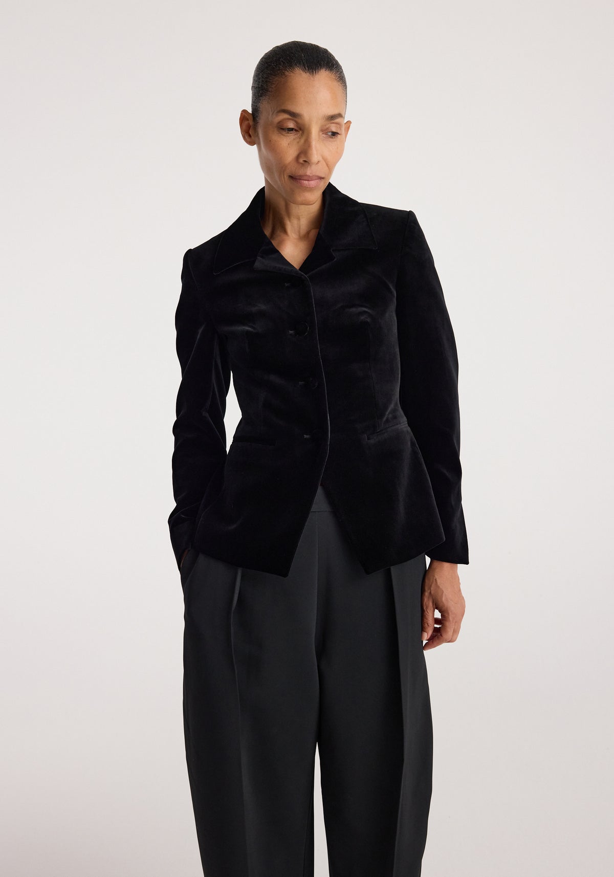 Woman wearing a Classic fitted velvet blazer | black with long sleeves and a tailored silhouette, paired with loose black pants against a plain background.