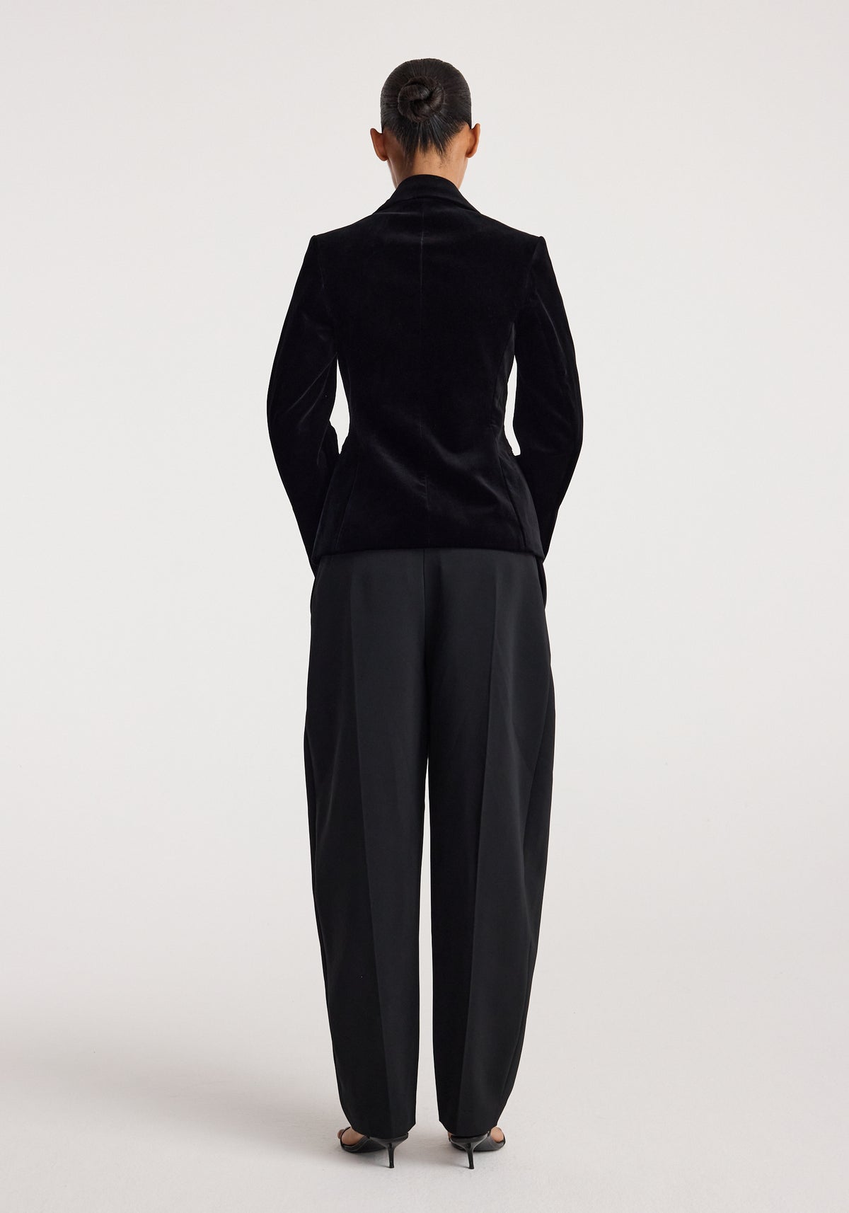 Woman seen from behind wearing a Classic fitted velvet blazer | black with tailored black pants and high-heeled shoes against a plain background.