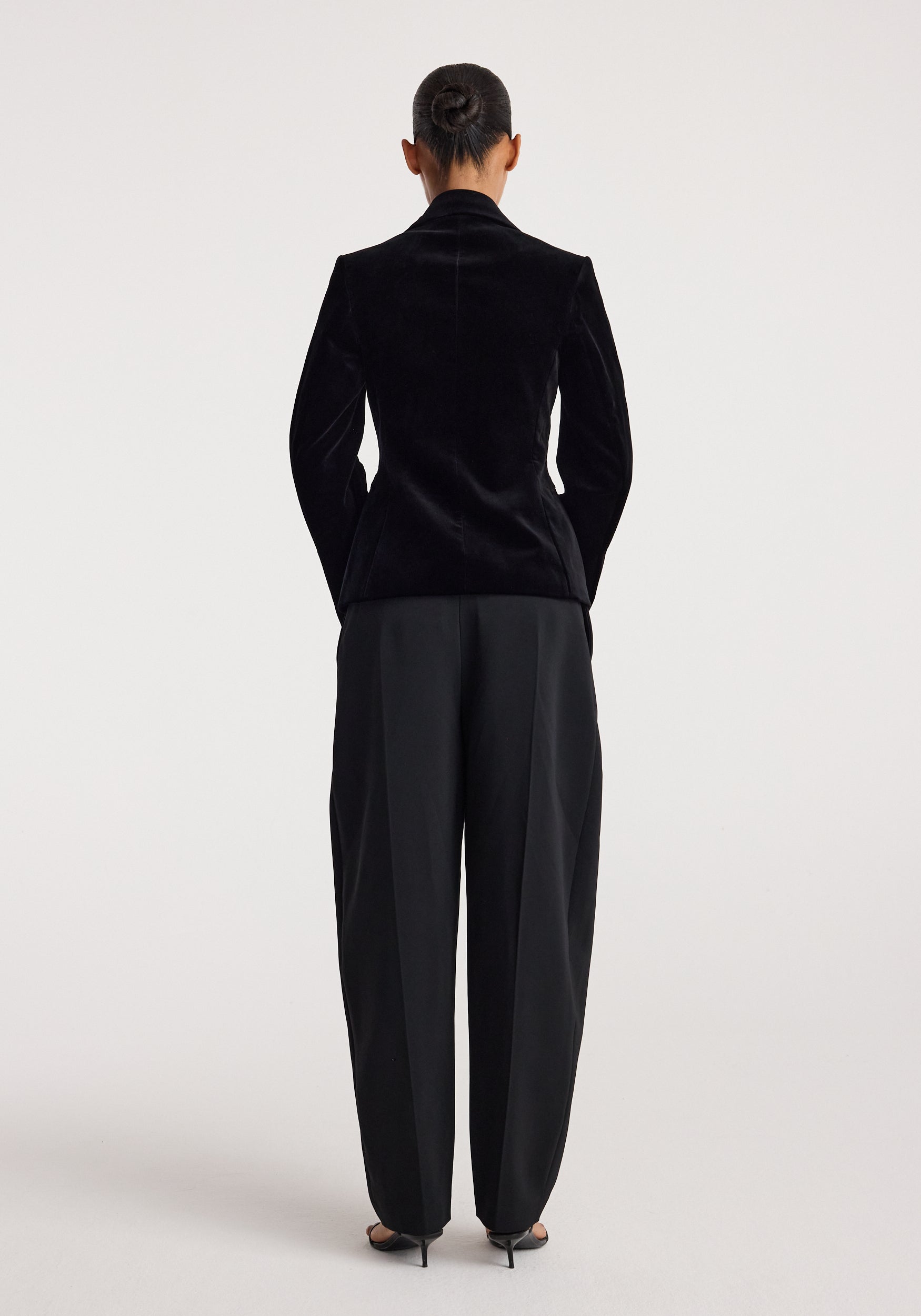 Woman seen from behind wearing a Classic fitted velvet blazer | black with tailored black pants and high-heeled shoes against a plain background.