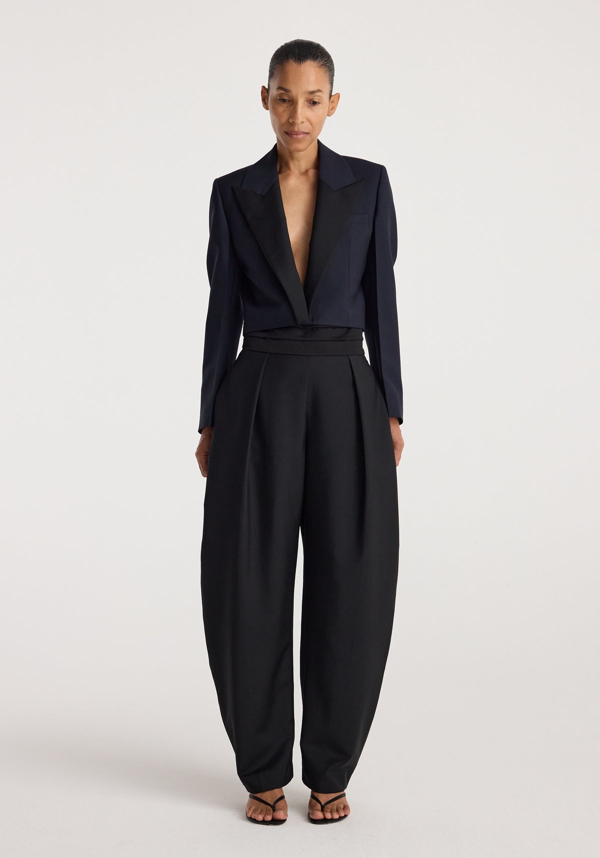Woman wearing a tailored Cropped blazer | navy with long sleeves and wide lapels paired with high-waisted black trousers and open-toe heels, standing against a plain background.